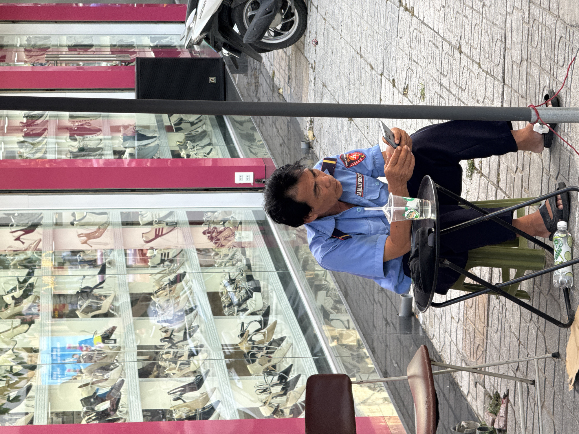 A man wearing a blue security guard uniform sits on a green stool next to a small black folding table, looking at his smartphone. A clear plastic cup with a green straw and a drink is on the table. Behind him, a shoe store's display windows are visible, filled with rows of women's shoes. A motorcycle is parked on the sidewalk to the right. The scene appears to be on a sidewalk in Nha Trang, Vietnam, suggested by the architecture and the style of the motorcycle.