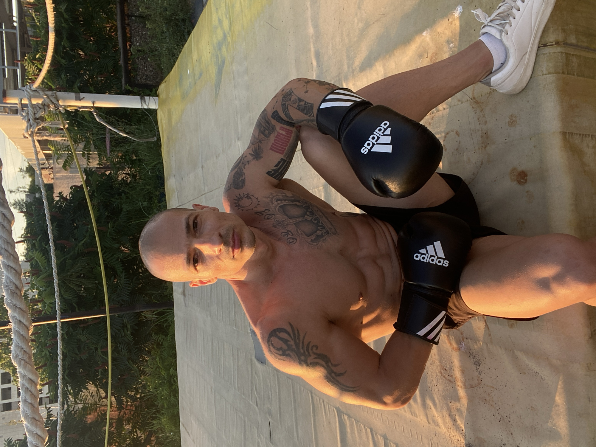 A bald, muscular man with numerous tattoos is sitting in a boxing ring. He is wearing black boxing gloves with white Adidas logos and black shorts. His left arm is tattooed with a tribal design, and his right arm and chest have various other tattoos, including the word "WORLD" in red lettering and a depiction of the sun. He is looking directly at the camera with a serious expression. The boxing ring appears to be outdoors, with ropes and a canvas floor visible. In the background, there are green bushes, a building, and a clear sky, suggesting an urban or semi-urban environment. The lighting suggests it is either early morning or late afternoon.