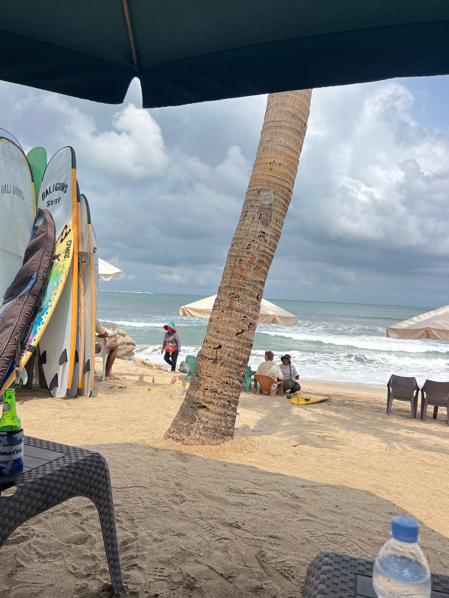 A beach scene in Kuta, Indonesia, under a cloudy sky features a collection of surfboards leaning against a palm tree. In the foreground, a woven black table holds a green bottle in a can koozie. Further back, two people sit on chairs facing the ocean, with a yellow surfboard placed on the sand nearby. A woman in a red hat and blue shirt walks along the shoreline. The ocean is visible with waves, and two umbrellas provide shade on the sand. The overall atmosphere suggests a relaxed beach setting.