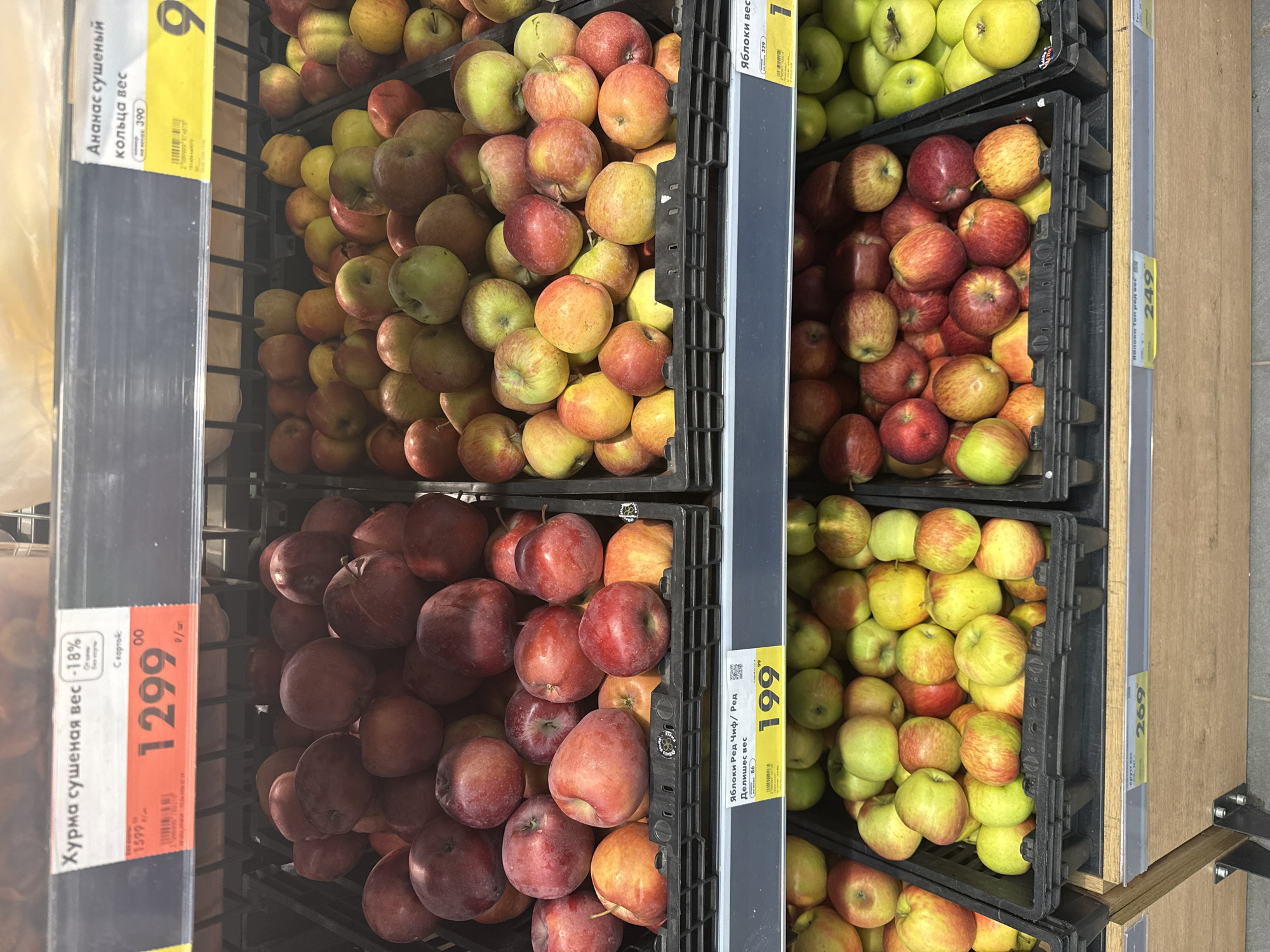 This is a photograph taken in a retail setting, most likely a grocery store, in Moscow, Russia.
The image shows a display of apples in stacked black plastic crates. There are several varieties of apples, ranging in color from green to yellow to red.
Price tags in Russian are visible next to the apples, indicating the prices for different varieties. Other items such as "Хурма сушеная вес," which translates to "Dried persimmon weight," and "Ананас сушеный кольца вес", "Dried pineapple rings weight" also have price tags.