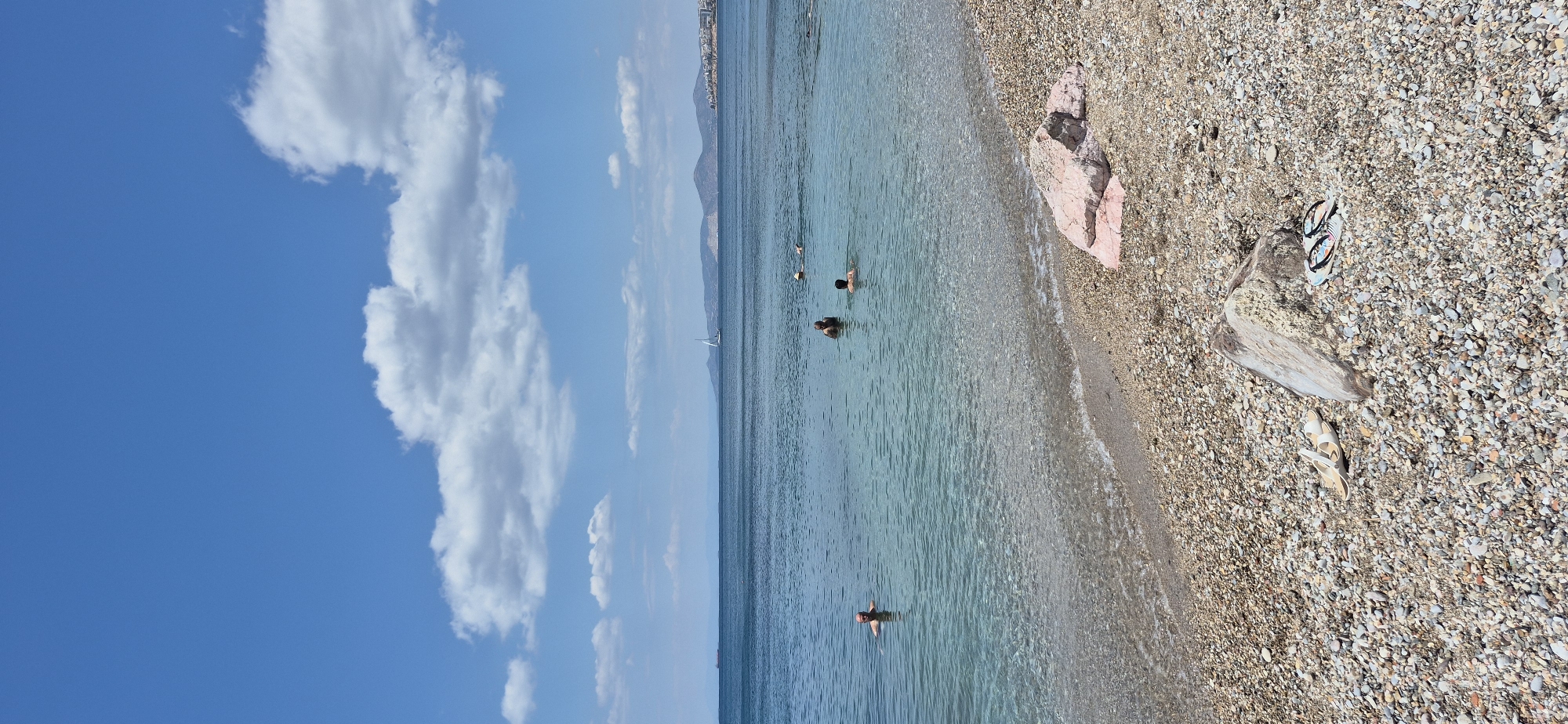 A wide shot captures a tranquil beach scene in Paleo Faliro, Greece. The foreground is a pebble beach with scattered rocks and a pair of white sandals and flip-flops. The clear, shallow water laps gently at the shore. Several people are visible enjoying the water, some swimming while others are partially submerged. In the middle ground, a sailboat glides across the calm sea. The background features a hazy coastline with hills and buildings, under a bright blue sky dotted with fluffy white clouds. A distant cargo ship is also visible on the horizon. The overall impression is one of a peaceful summer day at the seaside.