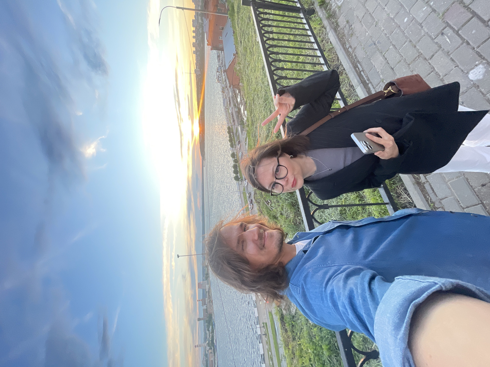 A selfie captures a man and a woman standing outside with a waterway in the background, likely taken in the evening, possibly at sunset. The man, positioned in the foreground, wears a blue denim shirt and has medium-length brown hair. He smiles slightly, looking at the camera. Behind him to the upper left, the woman wears glasses and a dark jacket, and she is giving a peace sign with her hand. She holds a smartphone. The background shows a body of water, with buildings along the shore. The sky, visible above the buildings, is illuminated with hues of yellow, orange, and blue, suggesting a sunset. A black metal railing is visible. The setting appears to be an urban area.