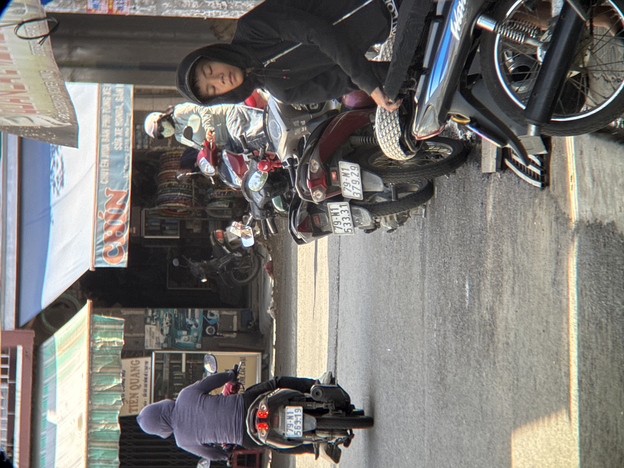 In Nha Trang, Vietnam, a street scene unfolds with multiple motorcycles and pedestrians. In the foreground, a young person in a black hoodie, with their eyes closed, leans on a motorcycle, appearing to rest. Behind them, a line of parked motorcycles is visible, some with license plates displaying Vietnamese registration. One license plate reads "79-NT 533.31." Further down the street, a person on a scooter, wearing a light-colored jacket and a white helmet, is partially obscured. They are positioned in front of a storefront with awnings and signage. The storefront appears to be a business selling motorcycle parts, with tires stacked inside. Another scooter, with a license plate reading "79-NT 563-19," is visible from the rear, with its rider wearing a purple hoodie. The environment suggests a typical urban street in Vietnam, characterized by the presence of numerous motorcycles and bustling commercial activity.