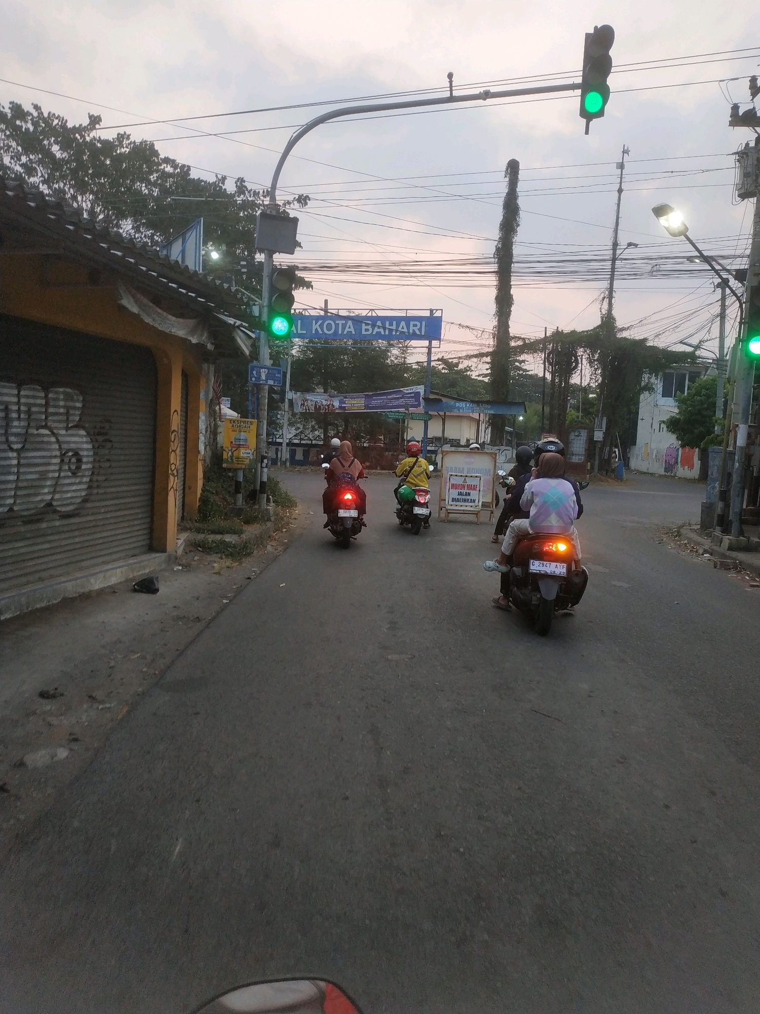 The image captures a street scene in Tegal, Indonesia, likely during the late afternoon or early evening due to the soft lighting. A traffic light is visible in the foreground, showing a green signal. Several people are riding motorcycles on the road. One motorcycle in the foreground has two occupants, the driver wearing a patterned jacket and the passenger a headscarf. Another motorcycle to the left carries a single rider in a yellow shirt, and further back, a motorcycle with two riders is also visible. The street is paved and appears to be an urban area, with buildings and trees lining the sides. A sign with Indonesian text, "AL KOTA BAHARI," is prominently displayed overhead. Graffiti can be seen on a shuttered storefront on the left side of the image. The overall atmosphere suggests a typical street in a Southeast Asian city, with a focus on daily commute and local infrastructure.