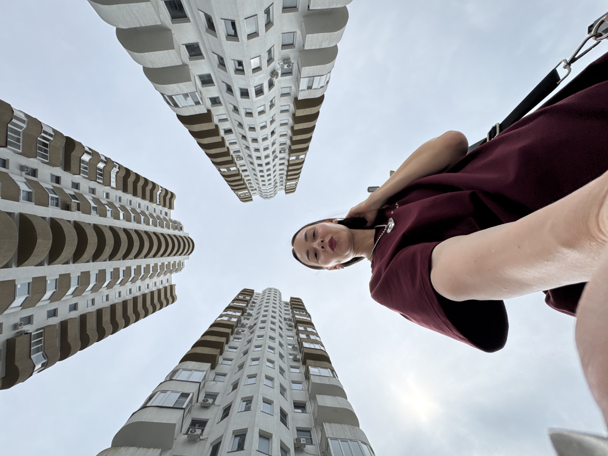 The image shows a low-angle, wide-shot selfie taken from below, with the camera pointing upwards towards the sky and surrounding buildings. The subject is a young woman with dark hair, wearing a maroon t-shirt and a necklace. She is touching her hair with her right hand, and her left arm is extended towards the camera, holding it. She has her lips pursed.
The background consists of four tall, modern apartment buildings, arranged in a symmetrical pattern around the woman, creating a sense of looking up from a central courtyard. The buildings have a distinctive architectural style, with curved balconies and alternating light and dark brown horizontal bands. The sky is overcast and pale grey, with a hint of sunlight peeking through from the left side of the frame.
The location is described as Sochi, Russia. The architectural style of the buildings and the cloudy sky are consistent with urban environments in Russia. The specific arrangement of the buildings suggests a residential complex or a similar development within the city. The composition of the image, with the woman positioned at the bottom and the buildings towering above her, emphasizes the scale and height of the architecture.