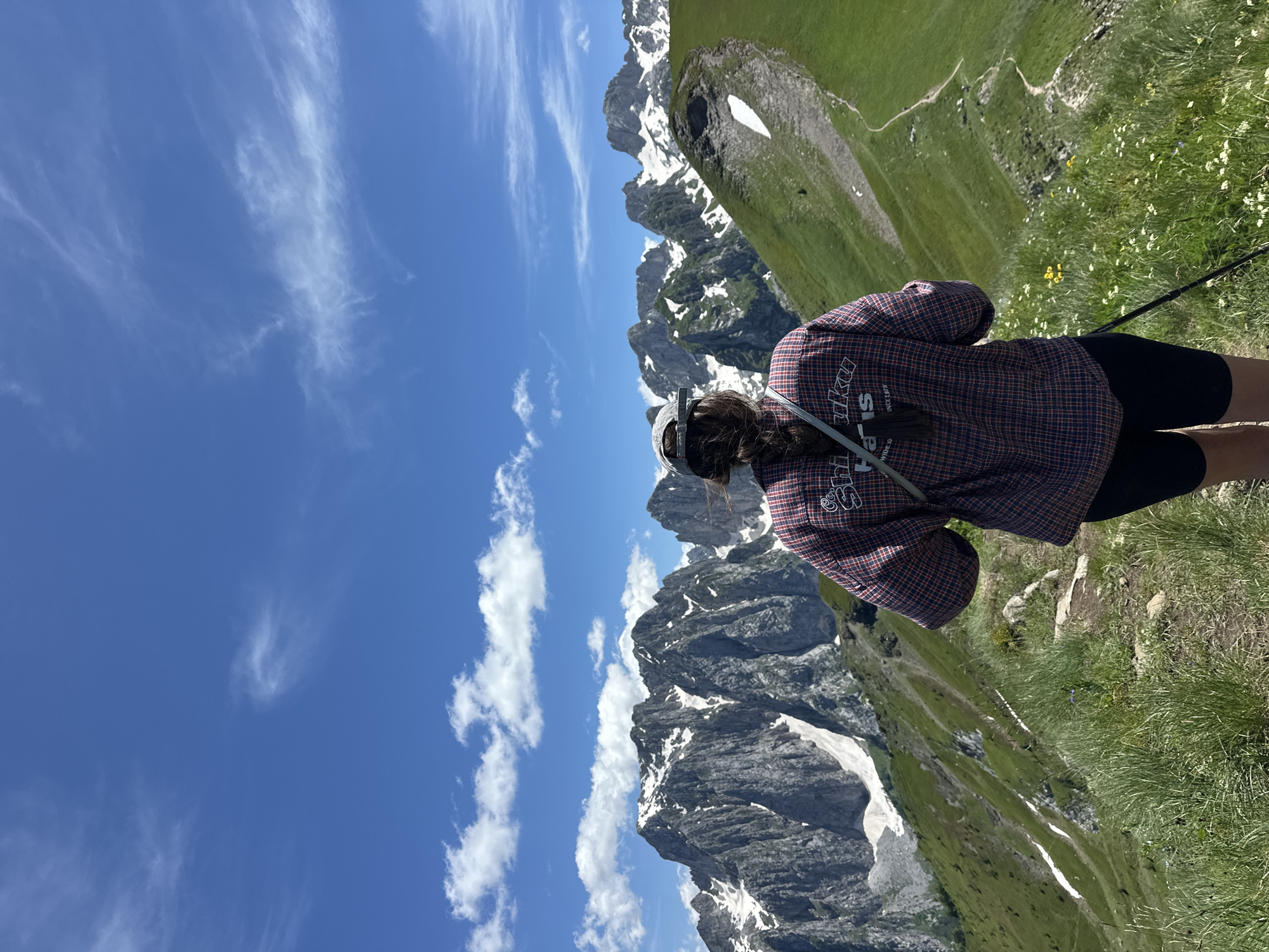 The image shows a person from behind, wearing a plaid shirt and a baseball cap, standing on a grassy mountain ridge. The person is holding a hiking pole. In the background, there are snow-capped mountains under a blue sky with scattered white clouds. The scene appears to be in a natural, mountainous environment, possibly a hiking trail. The location of Dolja, Montenegro is not depicted or suggested by the image content.