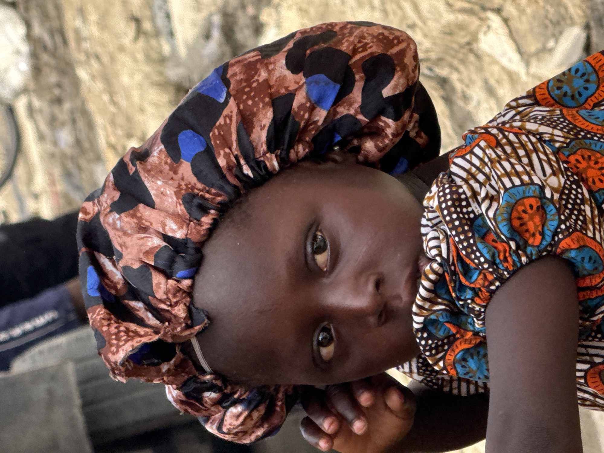 A close-up, tilted shot shows a young, dark-skinned person, likely a child, facing the camera directly. They are wearing a patterned head covering with brown, black, and blue designs. A colorful, patterned garment adorns their upper body, featuring orange, blue, white, and brown designs. The person rests their chin on their hand. The background appears to be a neutral, beige tone. The location is Ado, Nigeria.