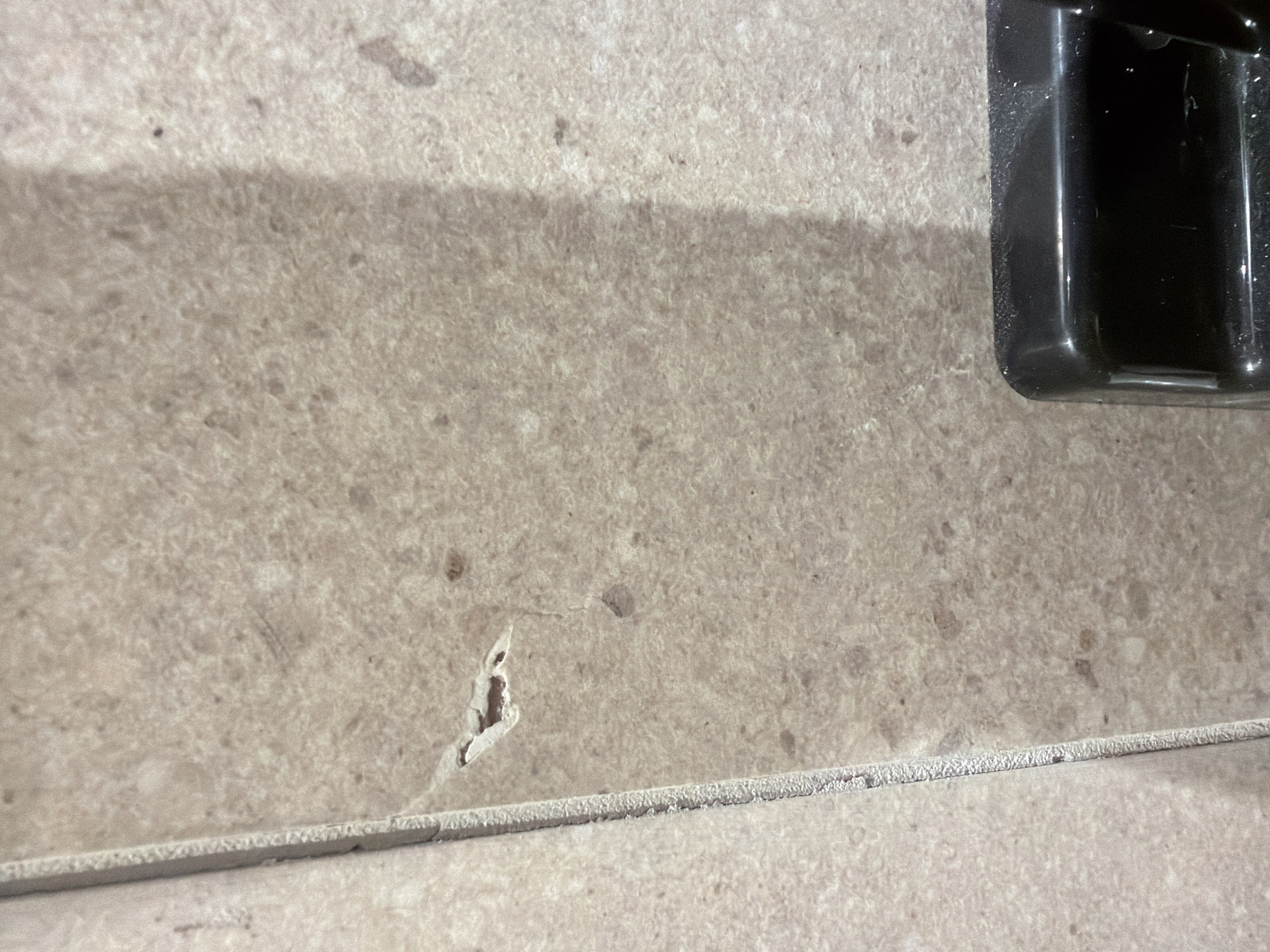 An overhead shot captures a section of tiled flooring. The tiles are a light brown with speckles of darker brown and beige. To the left, there is a clear grout line between two tiles, with the edge of the left tile showing a rough texture. In the upper portion of the image, there is a small chip or hole in the surface of one of the tiles. In the lower right corner, a black plastic object, possibly part of a vacuum cleaner or other appliance, is visible. The lighting suggests an indoor environment with a strong light source casting a shadow diagonally across the tiles. The scene appears to be in Jerusalem, Israel.