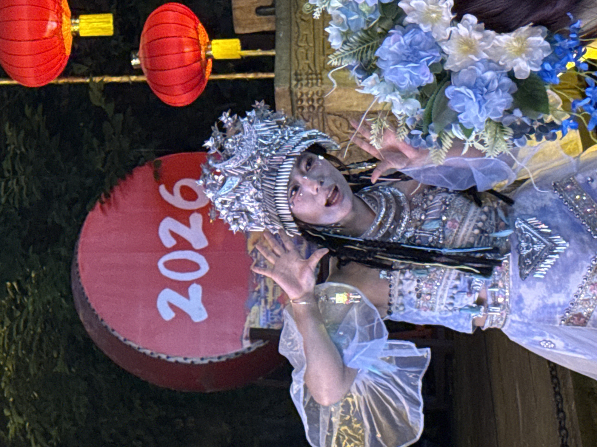 A photograph shows a person in an ornate costume and headdress performing in front of a red, circular sign that reads "2026." The costume is heavily embellished with sequins and intricate designs, and the headdress appears to be made of silver foil or a similar reflective material. The performer has dark hair and makeup, and is gesturing with both hands. Above and behind the sign are two red lanterns hanging from a string. The lighting in the scene appears to be artificial, giving a slightly staged or performance-like quality. The scene is located in Yuzhong District, China.