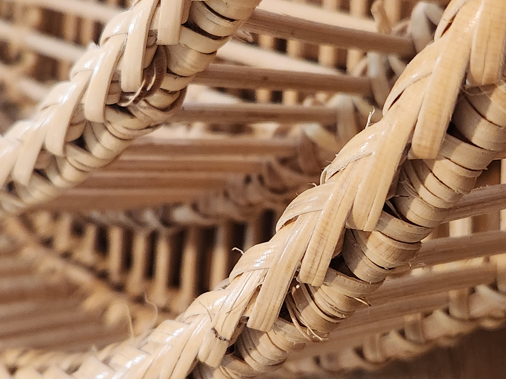 The image is a close-up, slightly angled shot of a woven wicker basket. The basket is constructed from thin, light-colored reeds that are tightly woven together. The texture of the reeds is visible, showing some variations in thickness and slight imperfections. The weaving pattern is intricate, with strands braided and layered to form the structure. Some parts of the weave are slightly frayed, suggesting age or use. The background is blurred and consists of more of the same wicker material, creating a shallow depth of field that focuses attention on the textured surface of the basket in the foreground. There are no people or distinct actions depicted. The setting does not provide any specific information to place it within the city of Maitland, United States of America.