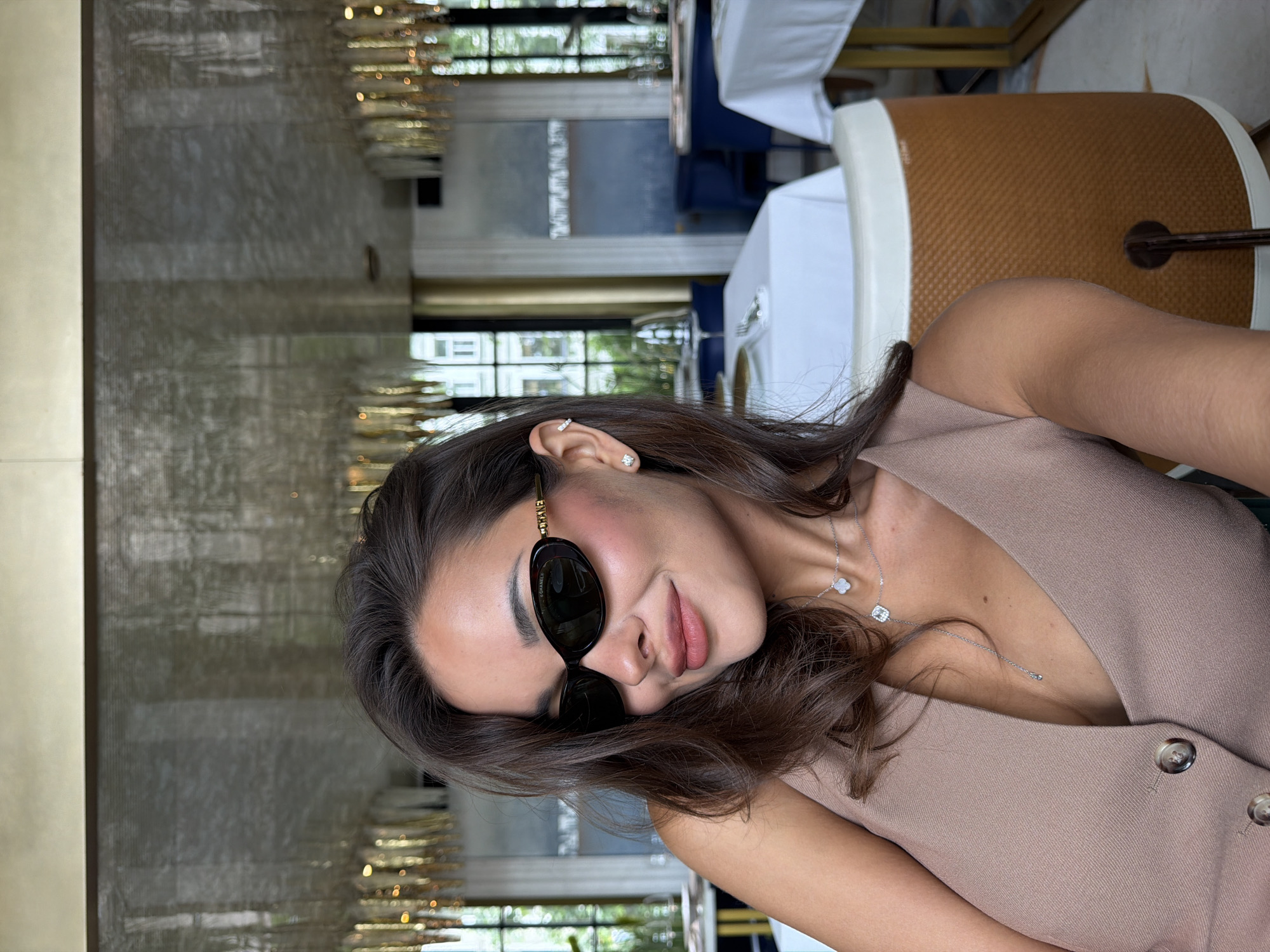 A young woman is depicted in a close-up, slightly tilted selfie or portrait, situated in what appears to be an upscale indoor restaurant or cafe in Moscow, Russia. She has dark brown hair and is seen with a gentle, soft smile, wearing dark, oval-shaped sunglasses that clearly display "CHANEL" text on the side of the frame. Her makeup includes a touch of blush on her cheeks and nude-pink lipstick.
She is adorned with multiple pieces of jewelry: a sparkling stud earring and a bar-shaped earring with small stones in her left ear. Around her neck, she wears two silver-colored necklaces; one features a distinctive four-leaf clover motif, and the other is a lariat-style necklace with a prominent square clear stone pendant. Her attire consists of a sleeveless, V-neck top or dress in a light brown or taupe color, fastened with two visible buttons on the front.
The background details an elegant setting. To her left, there's a textured wall, possibly marble or decorative paneling, reflecting ambient light. To her right, tables covered with white tablecloths and stylish chairs are visible. A chair in the immediate foreground features a unique brown woven pattern on its backrest with white trim. The scene is brightly lit, suggesting it is daytime, with light filtering from the background, creating a sophisticated and relaxed atmosphere.