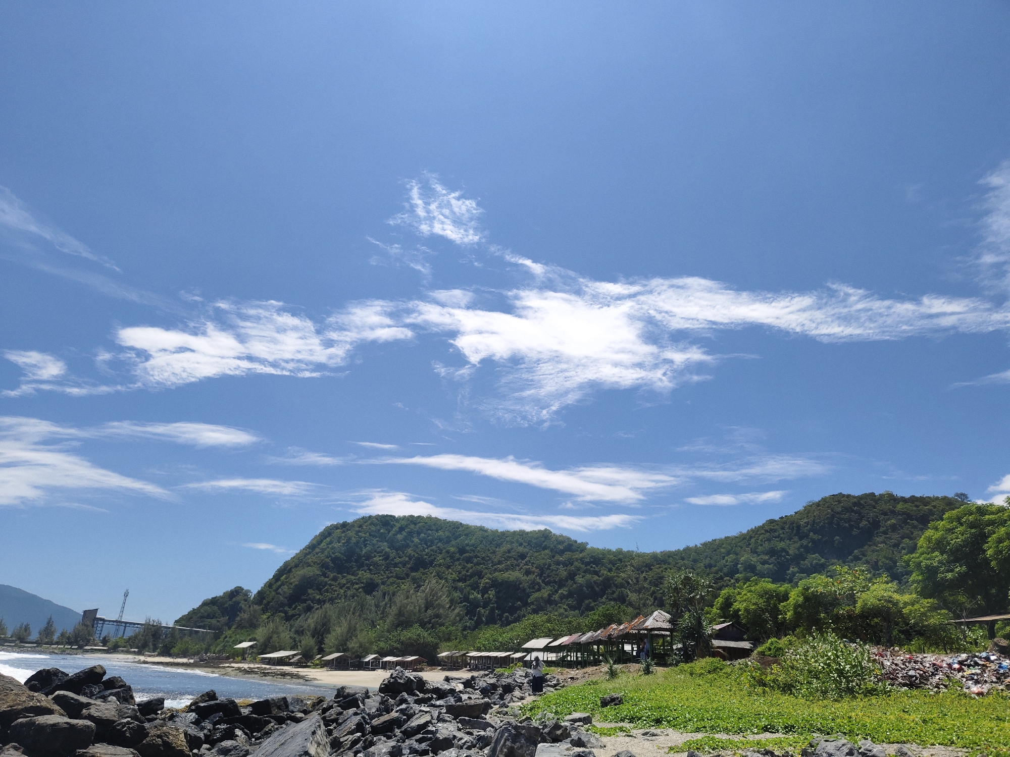 The image depicts a coastal scene in Naga Umbang, Indonesia. The foreground is dominated by a rocky shore with large, dark stones. Beyond the rocks, a sandy beach stretches along the coastline, with several small, rustic huts or shelters with thatched roofs situated on the sand. Some of these structures appear to be open-air, offering shade. A line of trees, including pine-like varieties and denser foliage, backs the beach. In the middle ground, a lush, green, heavily forested mountain rises dramatically, its slopes covered in dense vegetation. The sea is visible, with gentle waves lapping at the shore. In the far left of the image, a large industrial structure with a tall tower and connecting bridge-like elements stands near the coast. The sky above is a bright, clear blue with scattered white, wispy clouds. On the right side of the image, a pile of accumulated trash is visible amidst the greenery. There are no people clearly visible in the immediate foreground or middle ground, though a few figures can be discerned near one of the huts in the middle ground. The overall impression is a natural landscape with evidence of human presence and industrial development.
