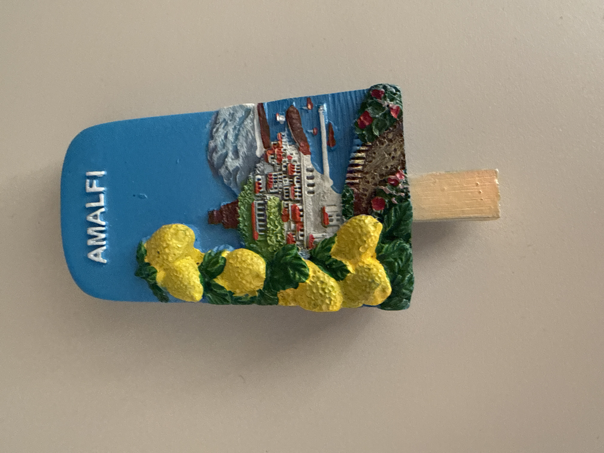 An embossed souvenir magnet, shaped like a popsicle, is prominently displayed against a plain, light gray background. The magnet depicts a vibrant, stylized scene of a coastal town, likely in a Mediterranean setting.
The main body of the magnet is a bright blue, representing either the sky or sea, with the word "AMALFI" written vertically in white, capitalized letters on the left side. The central part of the magnet features numerous white buildings with red roofs, characteristic of a town built into a hillside or cliff. These structures are clustered together, interspersed with green foliage. On the far right, there's a section of lush green vegetation adorned with small red flowers, possibly bougainvillea or roses, suggesting a warm climate.
The foreground of the magnet showcases a prominent border of sculpted yellow objects resembling lemons, complete with textured surfaces and green leaves. These lemons line the bottom edge of the scene, a common symbol of the Amalfi Coast. A light-colored, rectangular wooden piece, resembling a popsicle stick, protrudes horizontally from the right side of the magnet. The magnet has a three-dimensional, painted quality, with distinct textures for the lemons, foliage, and architectural elements. The lighting suggests a clear, bright environment, possibly daytime. The overall impression is that of a colorful, picturesque travel keepsake.
