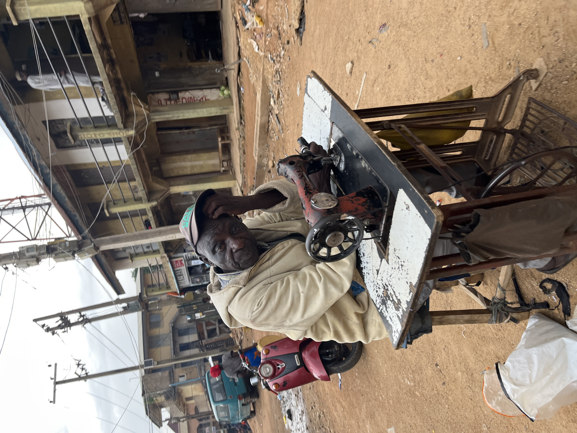 The image depicts a street scene in Jos, Nigeria. In the foreground, an elderly Black man wearing a light-colored hooded jacket and a baseball cap is seated at a vintage sewing machine. His right hand is resting on his forehead, and he appears tired or contemplative. The sewing machine is set up on a wooden table, which itself is supported by a metal frame with a foot pedal. The table surface is partially covered with what looks like scattered rice. Behind the man, a red scooter is parked. In the background, there are multi-story buildings with balconies and shops, typical of a commercial area. The street is unpaved and dusty, with scattered debris and electrical poles with numerous wires overhead. A turquoise van is visible in the mid-ground. The overall impression is of a busy, possibly impoverished, urban environment.