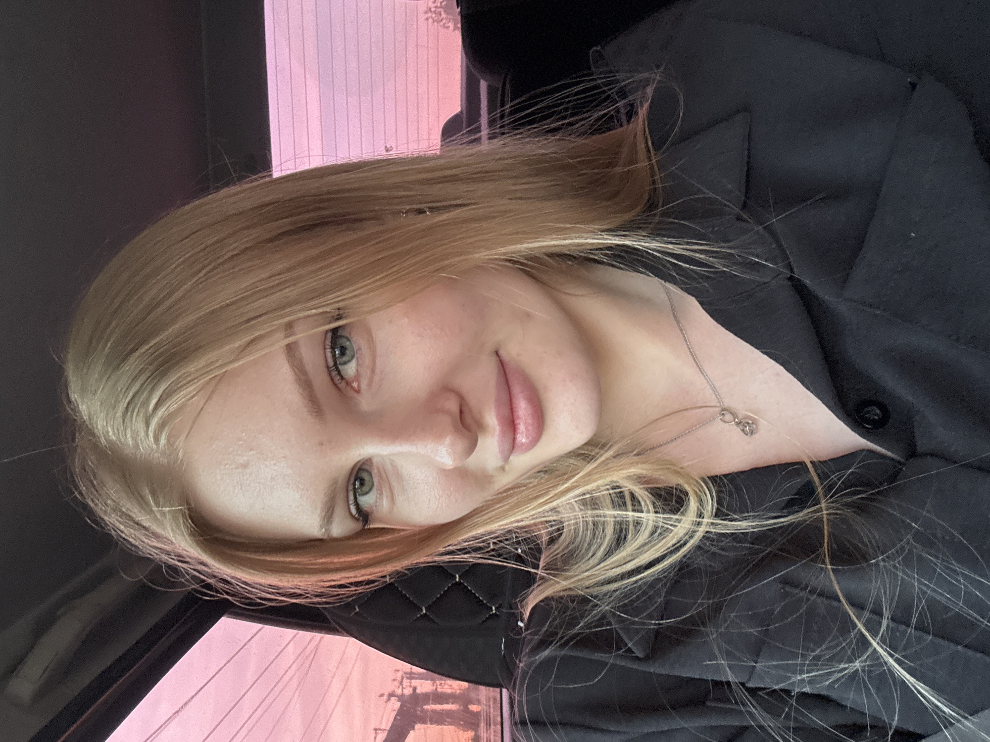 A blonde woman is pictured from the shoulders up inside a vehicle. She is wearing a black collared shirt and a silver necklace. She has fair skin, light blue eyes, and pink lips. The background visible through the car window suggests a sunset or twilight in the city of Кондратово, Russia. The interior of the car has a dark interior and a black quilted pattern on the headrest.