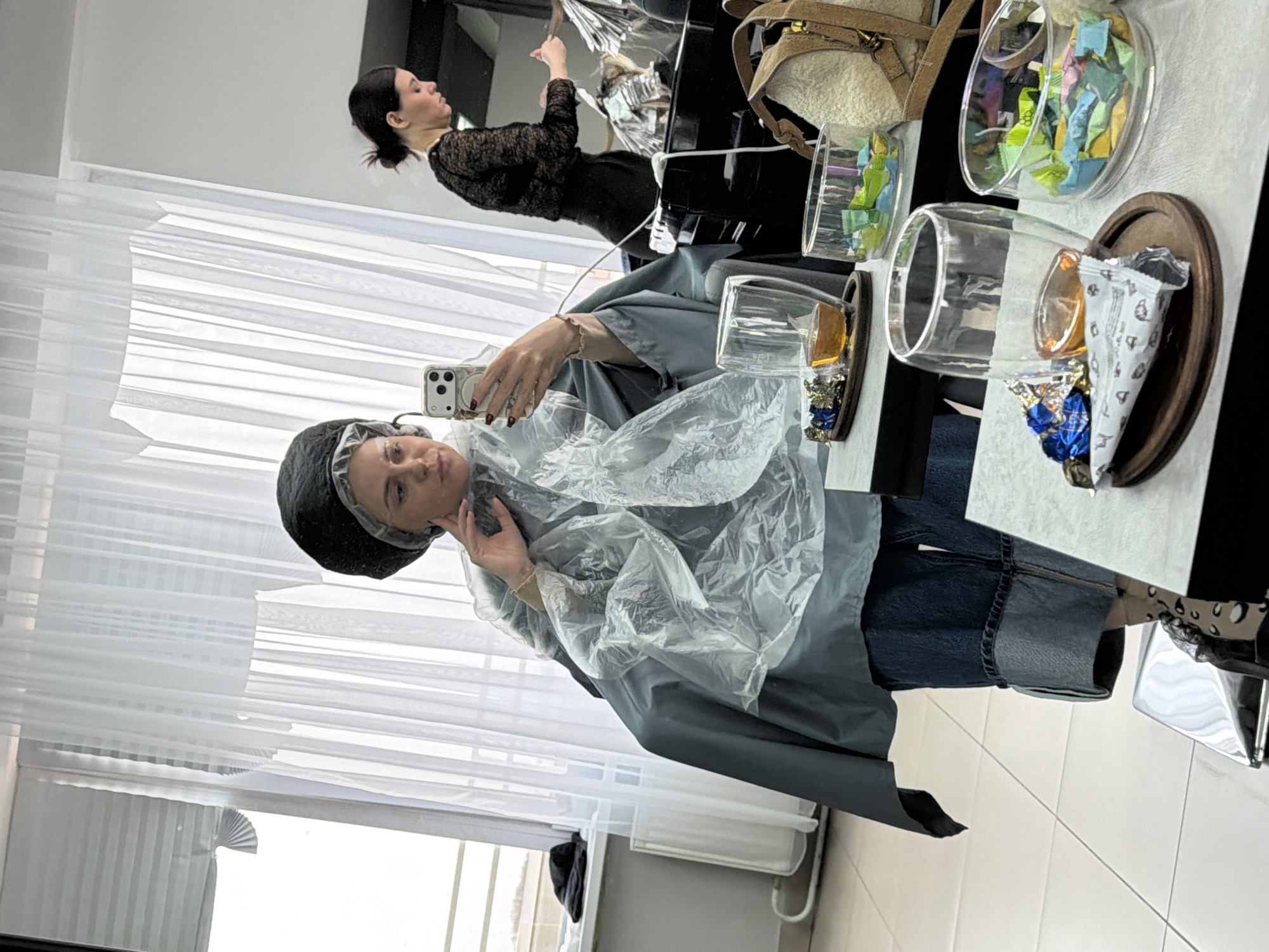 A woman wearing a black cap and plastic covering takes a selfie with a smartphone in what appears to be a hair salon in Votkinsk, Russia. The reflection in the mirror shows another woman in a black lace shirt working on a client's hair. Nearby are decorative clear containers on wooden bases holding items like candies and small objects, as well as a handbag. The background includes sheer white curtains and a radiator.