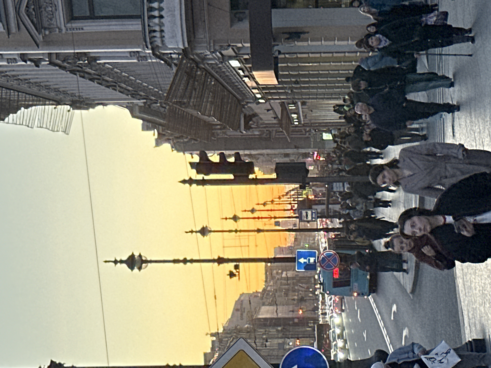 The image captures a street scene in Saint Petersburg, Russia, during what appears to be a late afternoon or early evening, indicated by the warm, golden hue in the sky. An avenue stretches into the distance, lined with ornate buildings. Regular intervals of street lamps line the thoroughfare. Pedestrians can be seen walking along the sidewalk. Street signs are visible among the cars and buses navigating the avenue.