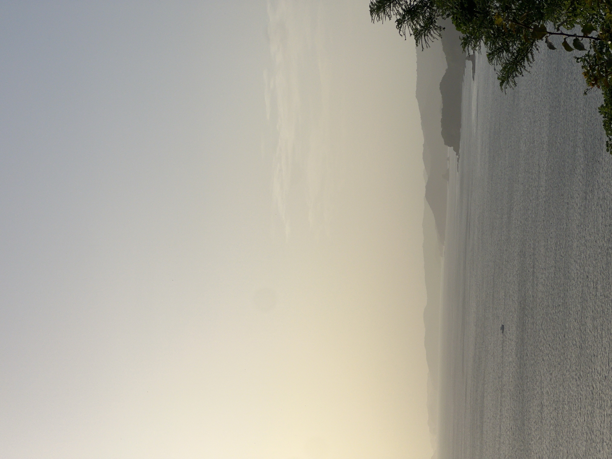 This is a medium-shot, vertical, eye-level, outdoor photograph taken from a high vantage point overlooking the Pacific Ocean in San Juan del Sur, Nicaragua. The sky occupies the top two-thirds of the frame, transitioning from a pale, hazy yellow at the bottom to a lighter, muted blue at the top. The ocean fills the bottom third, appearing dark gray with subtle ripples. In the middle ground, a series of layered, hazy green hills form a coastline. A single, dark-colored boat is visible on the water, slightly off-center to the left. In the foreground, on the right side of the image, the dark green foliage of trees partially obstructs the view of the ocean and coastline. The lighting suggests either early morning or late afternoon, with the sun likely low on the horizon, contributing to the hazy atmosphere. There are no people visible in the frame.