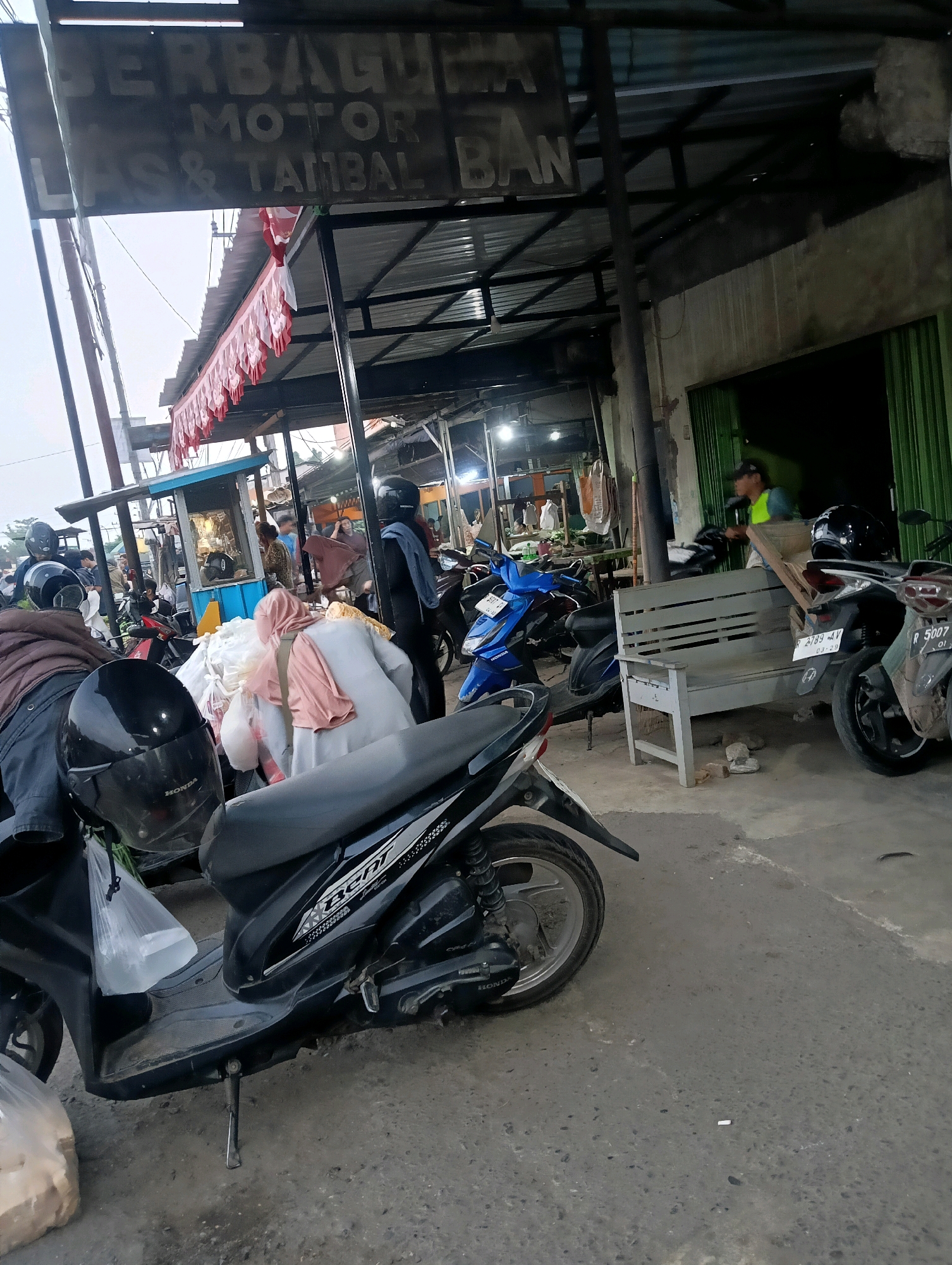The image depicts an outdoor scene in Purwareja, Indonesia, likely a street or a market area. A sign overhead reads "BERBAGAI MOTOR LAS & TAMBAL BAN," indicating a motorcycle repair shop specializing in welding and tire patching. Several motorcycles are parked in the foreground and background. In the immediate foreground, a black scooter is parked with its kickstand down. A black helmet rests on its handlebars. Further back, a blue motorcycle is visible. A light-colored bench is placed against a building, and a person in a neon green vest is standing nearby. In the middle ground, a stall with a blue facade is set up, with people interacting around it. Red and white decorative banners hang from an awning. The ground appears to be paved and somewhat worn. The overall impression is of a busy commercial area catering to motorcycle owners.