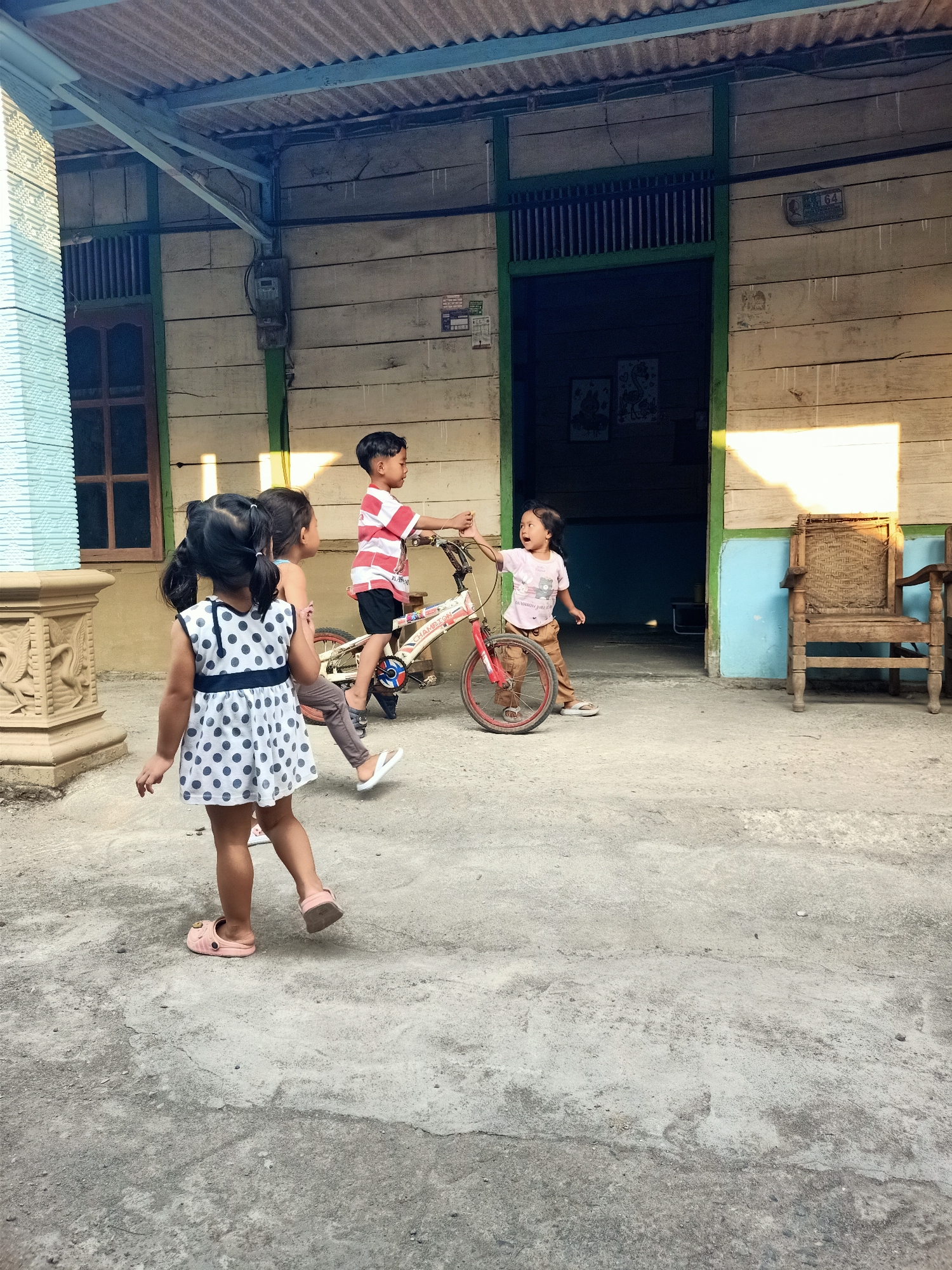 In the city of Kendung, Indonesia, on a concrete ground beneath a corrugated metal roof supported by a blue metal frame, a group of four young children are engaged in playful activity near a wooden building with a green-trimmed doorway. The building's exterior is constructed of horizontal light-colored wooden planks, and an electric meter is visible on the left side of the wall. To the right of the doorway, a woven wooden chair sits on the concrete.
In the foreground, a young girl wearing a white polka-dotted dress and pink sandals turns away from the camera, her dark hair in two pigtails. Behind her, another child, partially obscured, is also facing away.
Further into the scene, a young boy in a red and white striped shirt and black shorts stands on a red and white bicycle. He appears to be interacting with a girl in a pink t-shirt and brown pants who is reaching out with her right hand towards the boy and the bicycle. The girl in the pink shirt has her mouth open, as if speaking or laughing. The bicycle has a white frame with red accents and a red seat. The ground is a cracked and weathered concrete surface. To the left, a decorative light blue pillar with carved details stands prominently. The scene is illuminated by natural light, casting shadows on the ground and the building.