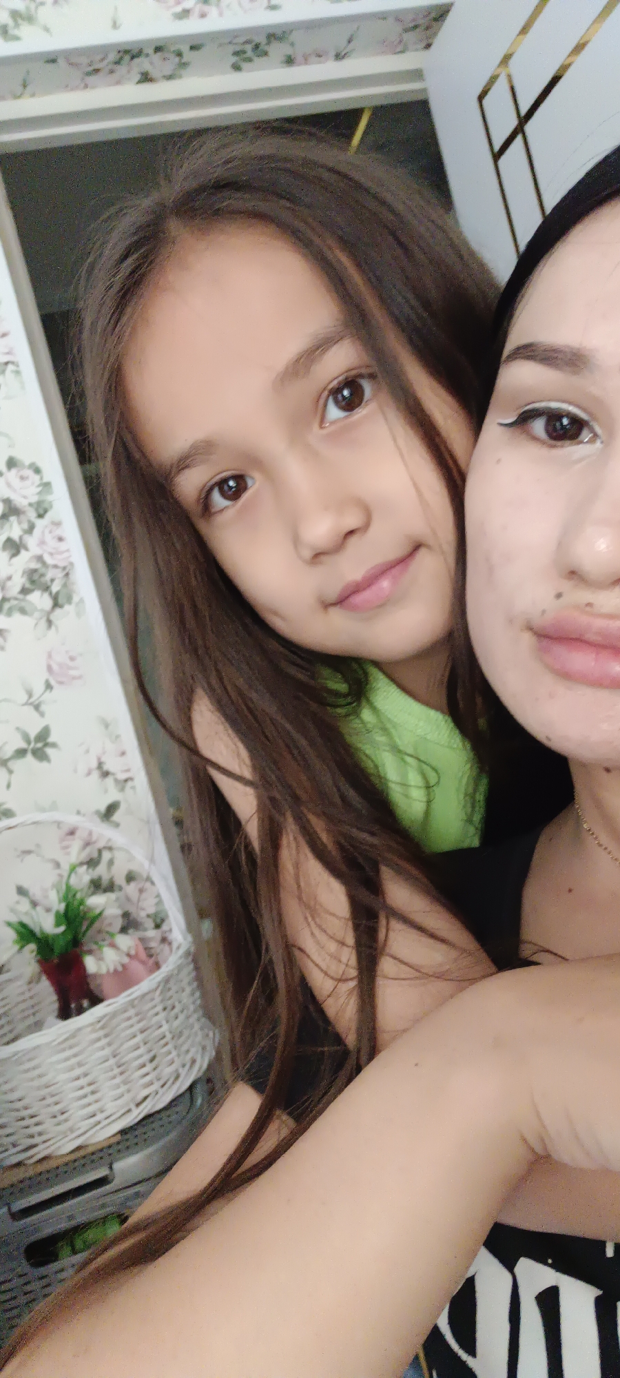 In this close-up selfie, a young girl with long, dark hair and brown eyes is positioned in front of a woman. The girl is wearing a bright green t-shirt and is looking towards the camera with a slight smile. The woman, partially visible, has her arm around the girl. She is wearing makeup, including white eyeliner, and a black top with a white gothic-style design on it. A portion of a white woven basket with artificial flowers is visible in the lower left corner of the frame, against a wall with a floral wallpaper pattern. The overall lighting suggests it is daytime. The image appears to be taken indoors, likely in a home setting.