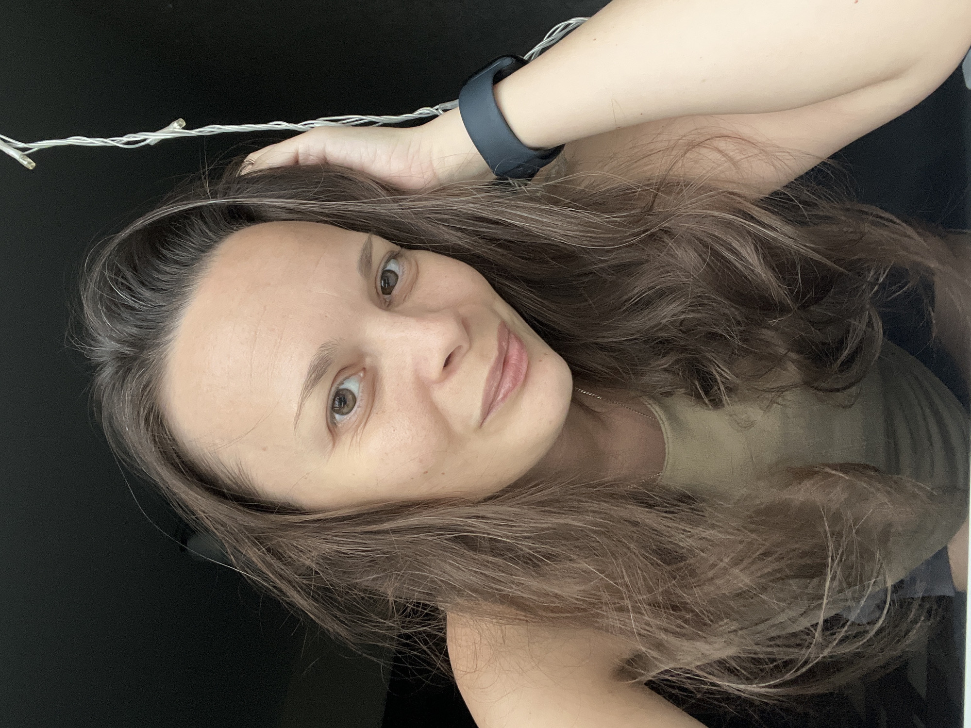 A woman is pictured in a close-up selfie. She has long, wavy brown hair and is looking towards the camera with a slight smile. She is wearing a light-colored tank top and a dark smartwatch on her left wrist. Her right hand is raised and resting on her head. Behind her, the background is dark and out of focus, with a string of small lights visible to her right. The image does not contain any information about the location in Tolyatti, Russia.