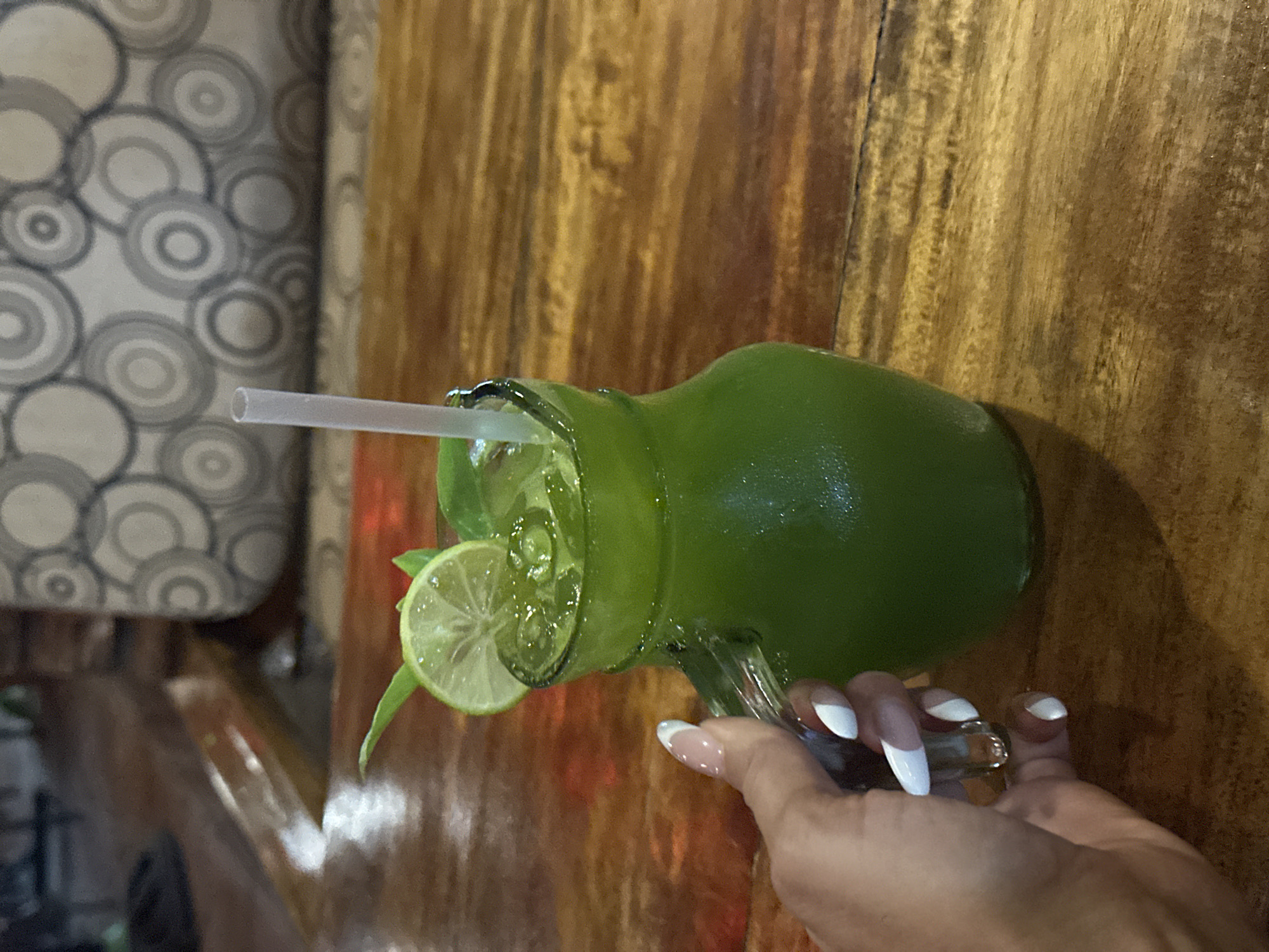 In Phu Thuy Ward, Vietnam, a hand with white-tipped French manicured nails is holding a clear glass pitcher filled with a bright green beverage. The pitcher contains ice and is garnished with a lime slice and a sprig of greenery. A white straw extends from the drink. The pitcher rests on a wooden table, and in the background, a cushioned bench with a circular pattern is visible.