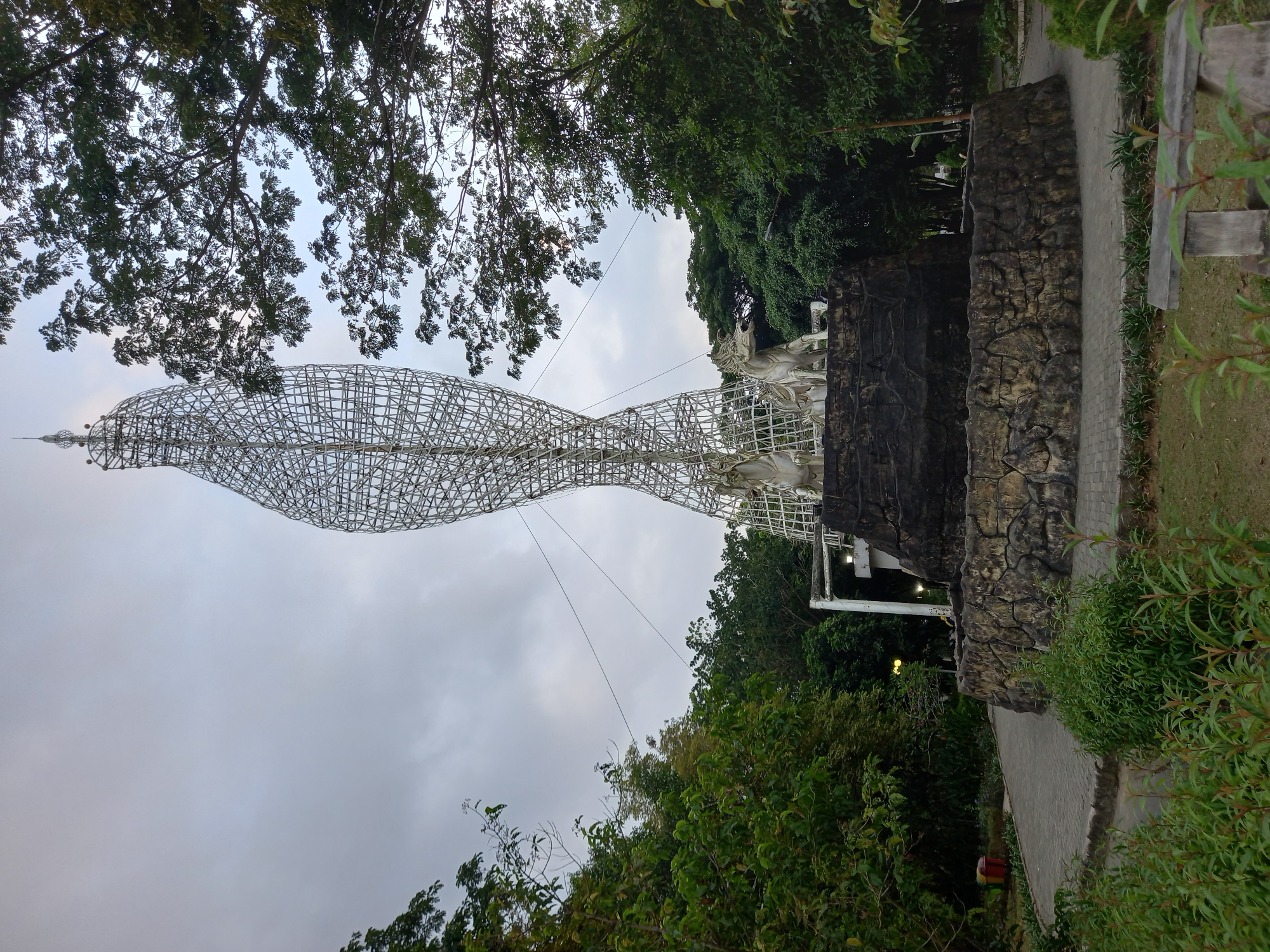 The image shows the Mahakam Lampion, a landmark in Samarinda, Indonesia. The sculpture is a tall, twisted metal structure resembling a flame, with two white horse statues at its base. The sculpture is surrounded by lush green trees and a paved pathway. The sky is overcast, suggesting it might be a cloudy day. The overall scene appears to be a public park or garden, likely a popular spot for visitors to the city.