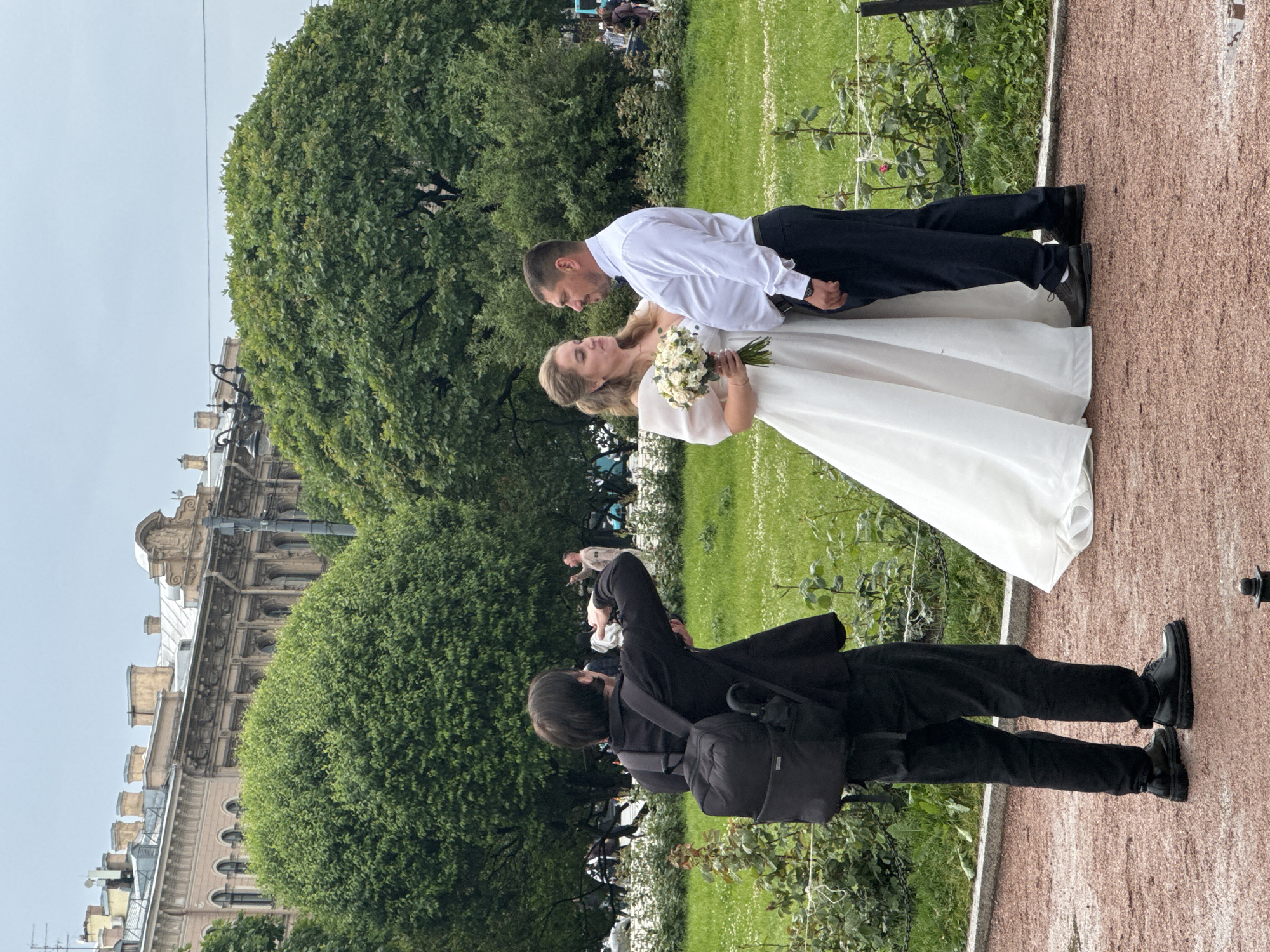 A wedding photographer, seen from behind wearing a black jacket and backpack, photographs a wedding couple in a park in Saint Petersburg, Russia. The bride wears a white wedding dress and holds a bouquet of white flowers, while the groom wears a white dress shirt and dark trousers. They stand in front of lush green trees, with a historic building visible in the background. The ground is a mixture of gravel and grass.
