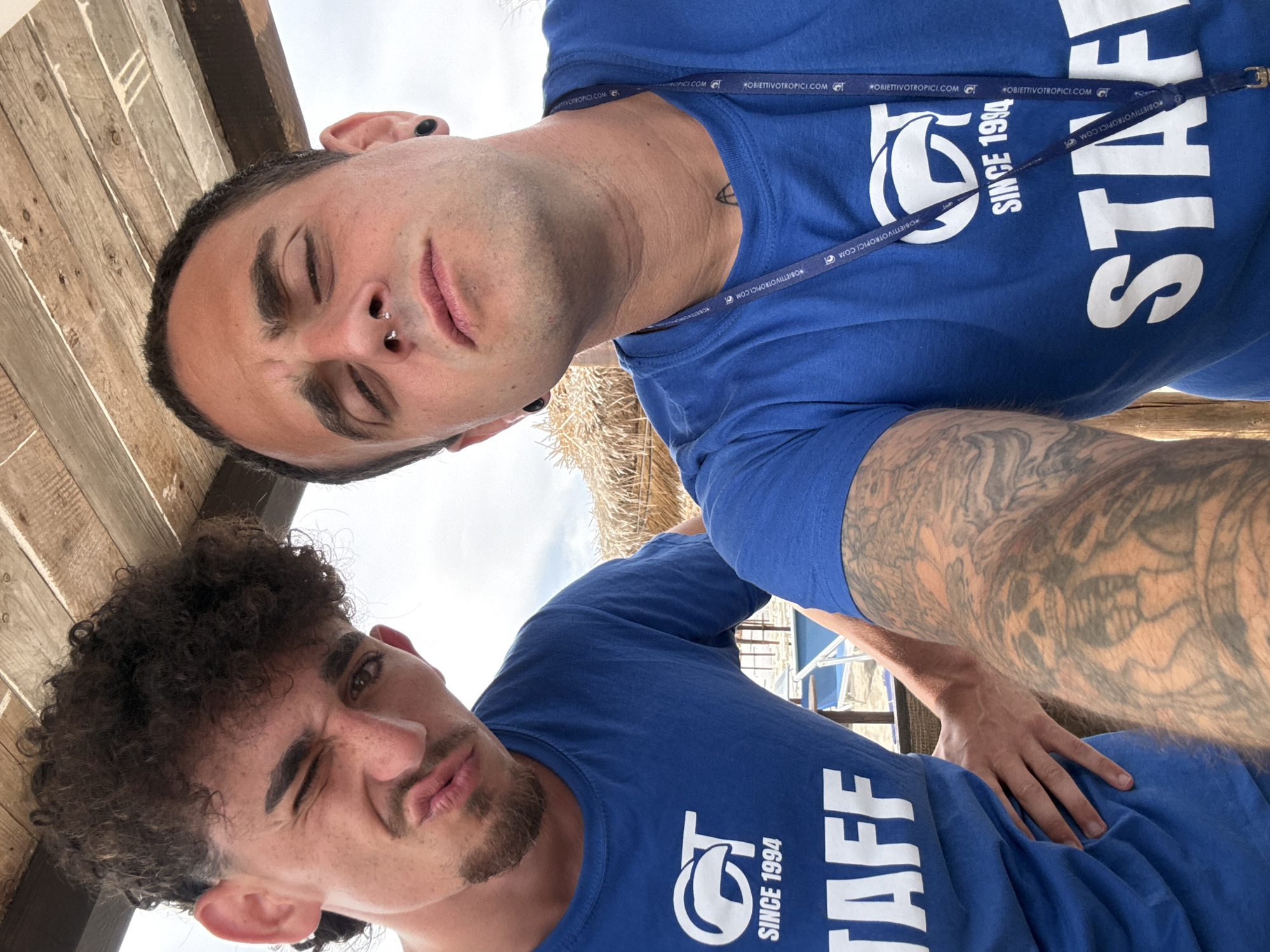 Two young men, likely staff members, are taking a selfie outdoors under a wooden structure. The man on the left has curly brown hair, a goatee, and is winking, with his mouth pursed. He is wearing a blue t-shirt with a white logo that says "GT SINCE 1994 STAFF." The man on the right has dark hair and a septum piercing. He is wearing a similar blue t-shirt with the same logo and "STAFF" printed on it, and a lanyard around his neck. His right arm, which is tattooed, is resting on his leg. The background shows a beach with lounge chairs and a thatched umbrella. The overall setting suggests a resort or beach club environment.