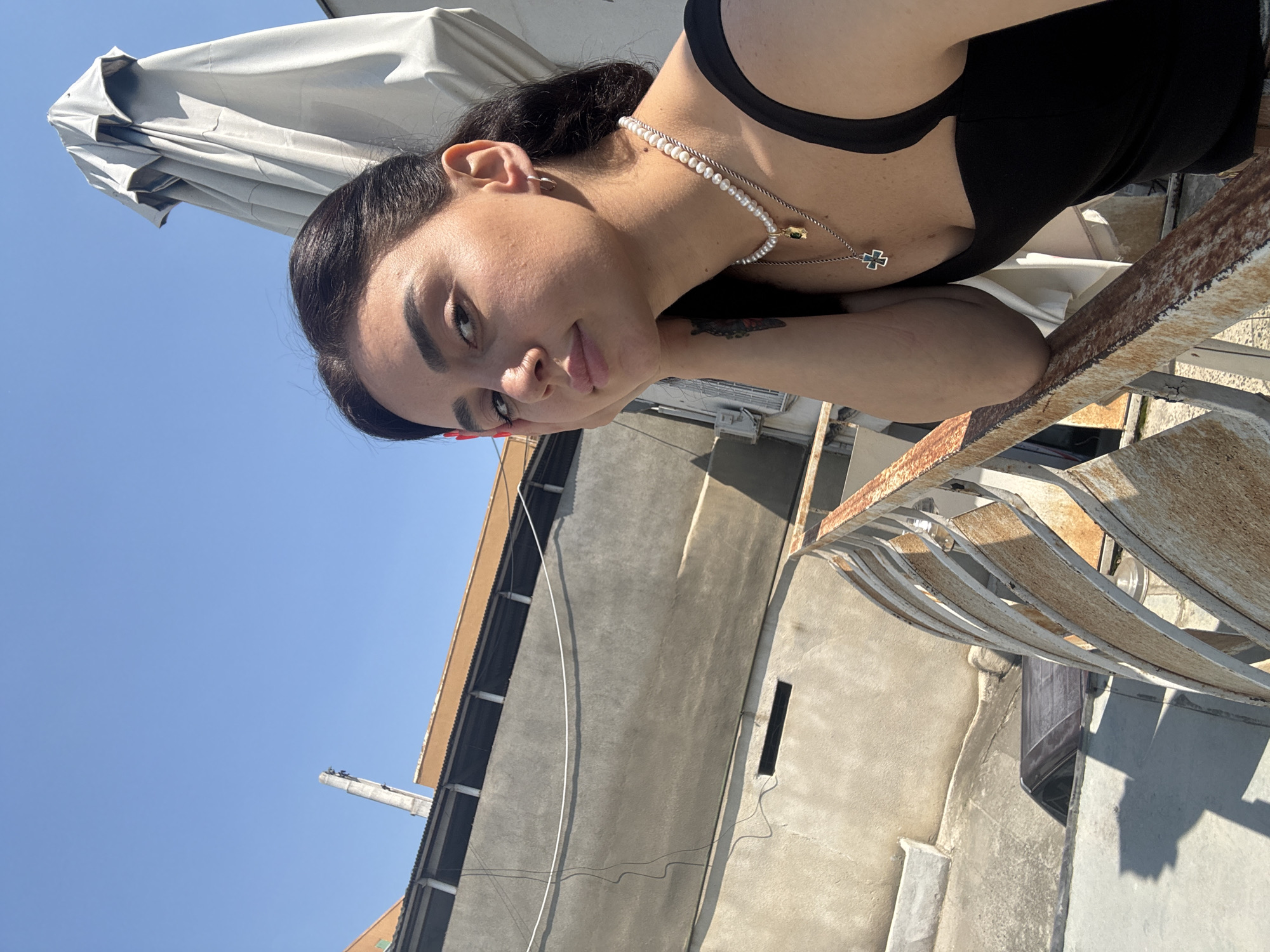 A young woman with dark hair and a thoughtful expression rests her chin on her hand as she poses for a selfie on a rooftop in Tbilisi, Georgia. She is wearing a black tank top and two necklaces, one of which is a pearl choker and the other a silver chain with a cross pendant. The background shows a clear blue sky, a gray building with an orange trim, a tall metal pole, and a large, folded umbrella. A rusty metal railing runs along the edge of the rooftop in the foreground, casting a shadow on the ground.