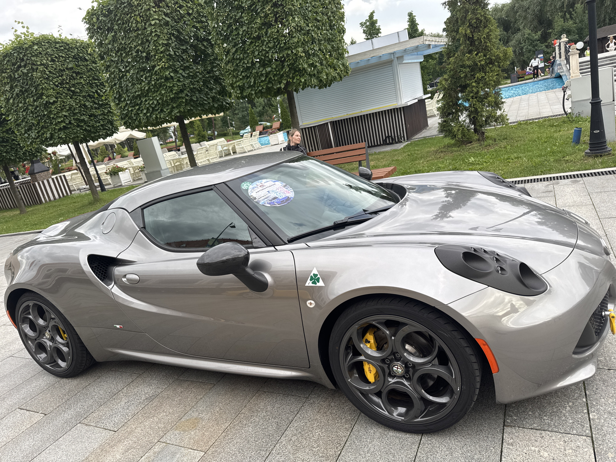 This media file features a silver Alfa Romeo 4C sports car parked on a paved surface. The car is the primary object in the foreground, with its distinctive design and a green cloverleaf decal on the fender. A woman is partially visible sitting on a wooden bench behind the car, looking to her left.
The scene appears to be an outdoor recreational area, possibly a park or a venue with a swimming pool in the background. The pool area has blue water and is surrounded by a paved deck. Several lounge chairs and umbrellas are visible, suggesting a place for relaxation or a resort-like setting. In the background, there are trees, some of which are neatly trimmed into geometric shapes, and a white building with a striped facade, possibly a cafe or a kiosk.
The weather appears to be overcast, indicated by the diffused light and the gray sky. The time of day is likely daytime. The presence of the sports car, the woman on the bench, and the general outdoor setting suggest a casual event or a public gathering, possibly an exhibition or a social outing. Some text is visible on a sticker on the car's windshield, which seems to be a circular decal with multiple small images and text in Russian. The overall mood of the scene is relaxed and leisurely.