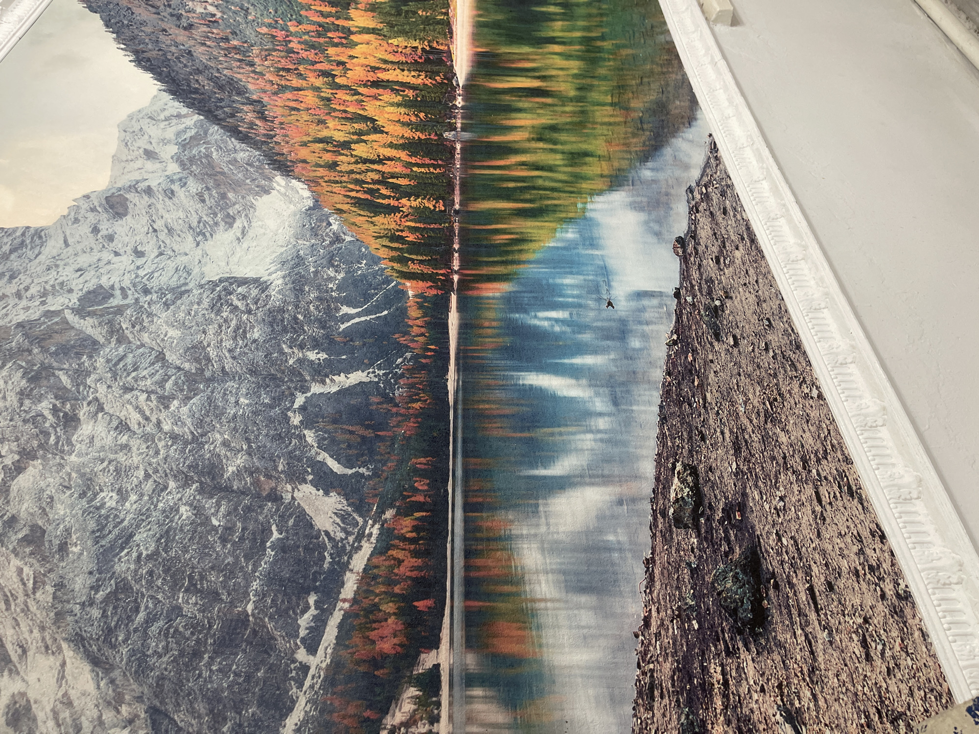 This media file shows a vertically oriented photographic print of a mountainous landscape scene with a reflection in a body of water. The print is framed by a white, ornate border. On the left side of the print, a snow-capped mountain is visible. To the right of the mountain, a forest with trees in autumn colors of orange and yellow lines the edge of a calm body of water. The water reflects the trees and the sky. The right side of the print depicts a rocky surface, possibly the shore or another part of the landscape, with some greenery or moss growing on it. The scene appears to be from daytime, with a clear sky suggested by the reflections in the water. There is no text visible in the image. The location context provided is Bishkek City, Kyrgyzstan.