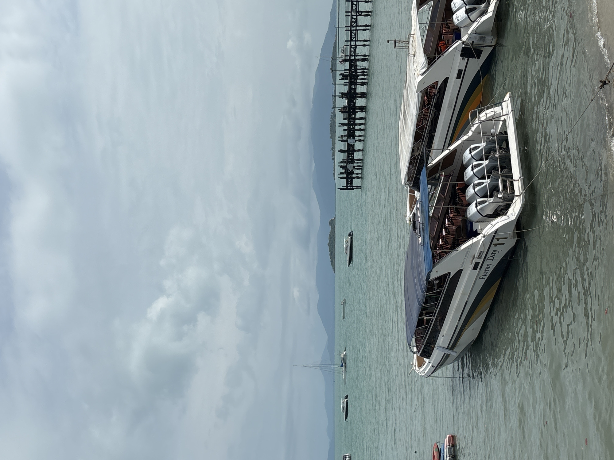 The image shows a coastal scene in Baan Bang Rak, Thailand, under a cloudy sky. Two speedboats are docked near the shore; one is labeled "Funny Day 11". A wooden pier extends into the water. Several other boats and a sailboat are visible in the distance. Mountains are present on the horizon.