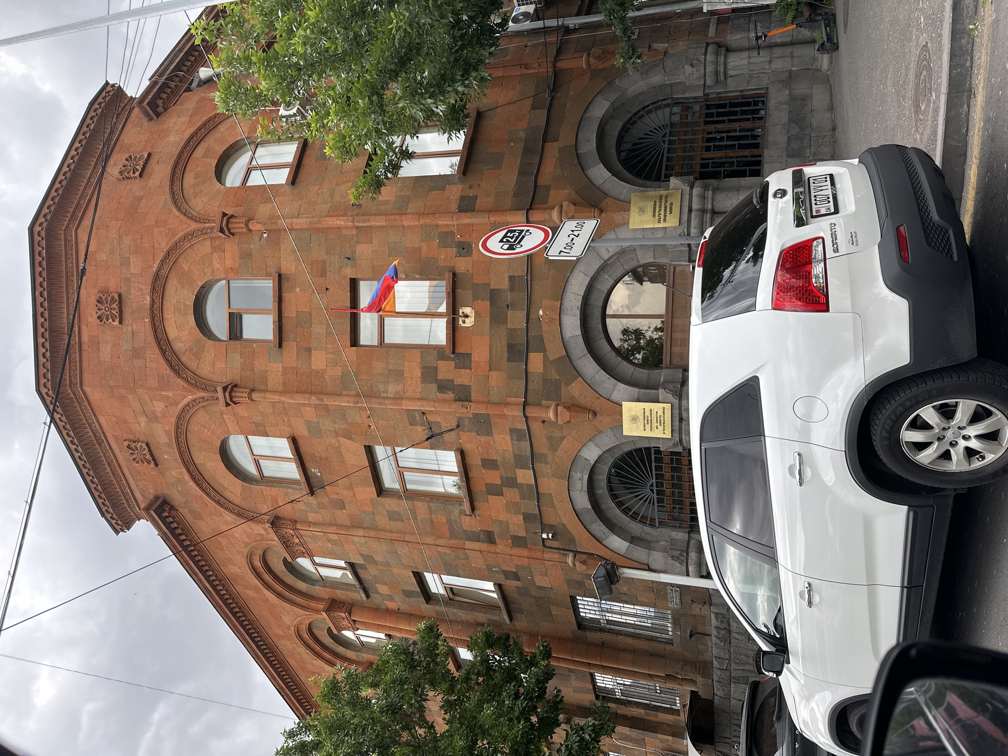 The image shows a building in Yerevan, Armenia, with a white Kia Sorento parked in front. The building is made of red and brown stone and has several arched windows. An Armenian flag is displayed in one of the windows. Road signs indicate a weight restriction and a time restriction from 7:00 to 21:00. Signage is visible on the building in Armenian. The building’s architecture is distinctive, with intricate stonework around the windows and along the roofline. The vehicle has a license plate of "00TYY001," which can be seen on the back of the car. The sky is overcast. Tree branches and the car's mirror partially obscure the foreground. A street sign is seen on the side of the building. The focus is on the building and the vehicle parked in front of it.