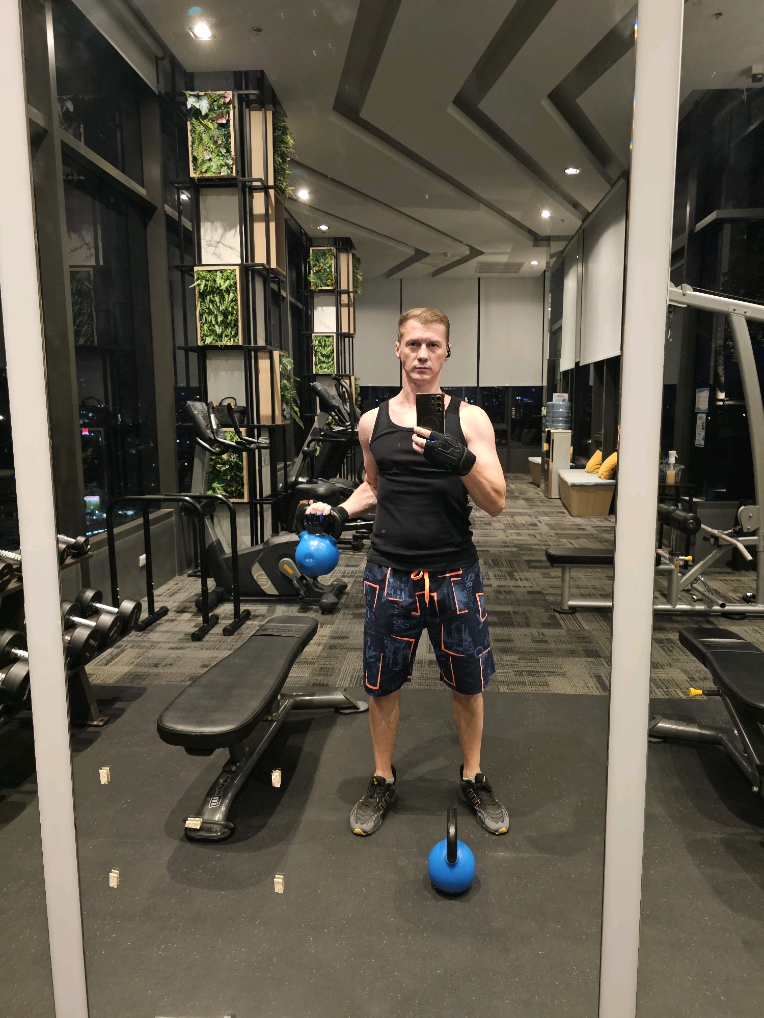 A fair-skinned man in his 40s stands in a gym, taking a mirror selfie. He is wearing a black tank top, patterned shorts, black gloves, and grey athletic shoes. He is holding a blue kettlebell in his right hand. Another blue kettlebell is on the floor in front of him. The gym appears to be modern, with gym equipment, benches, and large windows overlooking a city at night, possibly Bangkok, Thailand. The gym features decorative vertical planters with greenery.