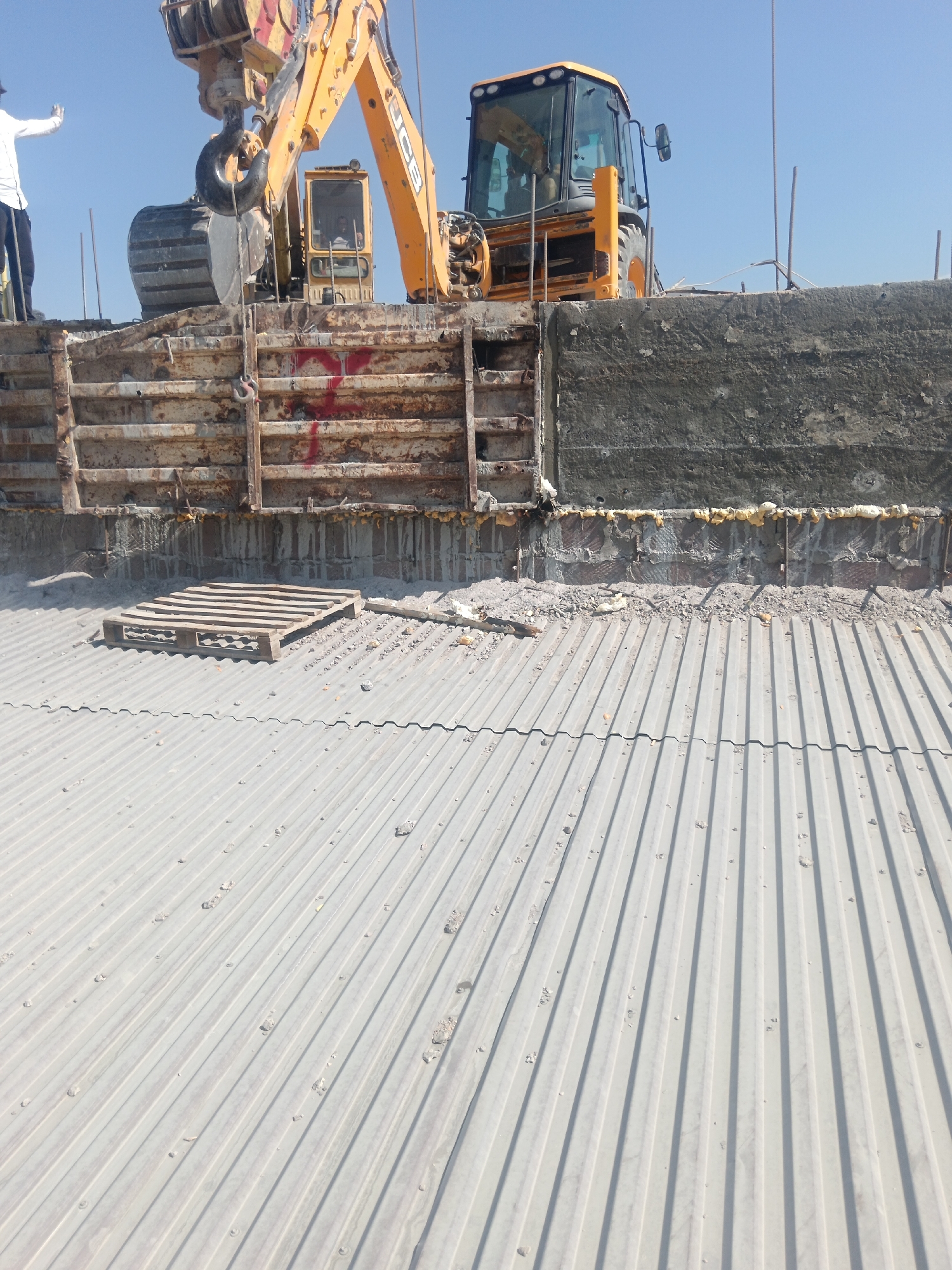 A construction site is depicted in Aragatsavan, Armenia, under a clear blue sky. In the background, a yellow JCB excavator is positioned, with a person operating it visible through the cab window. In the foreground, a person in a white shirt and dark pants stands on a construction platform, raising their left arm. The scene shows partially constructed walls with visible rebar and concrete, and a corrugated metal roof in the lower portion of the image. A wooden pallet rests on the metal roof. The overall impression is of ongoing construction activity.