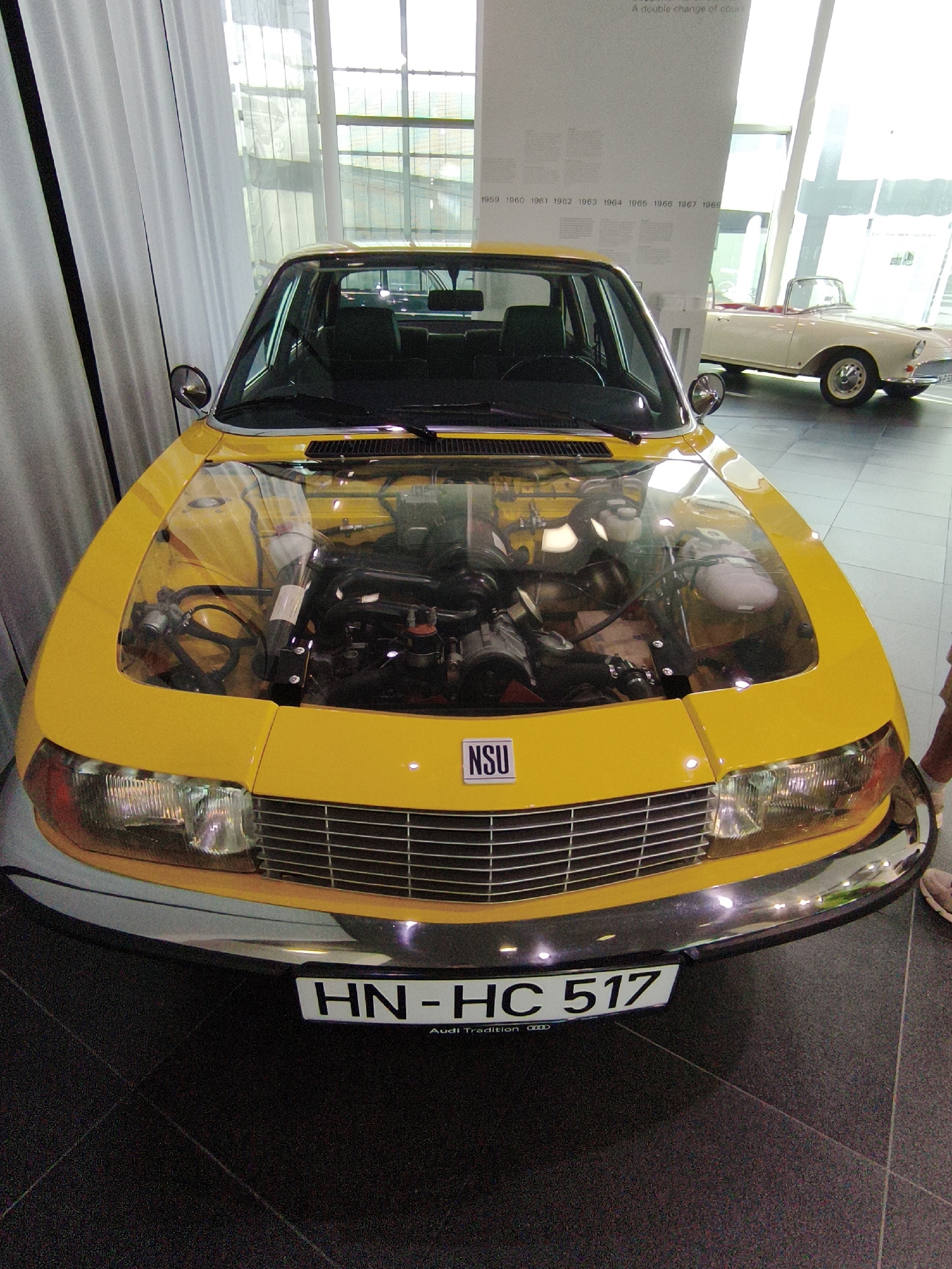 The image shows a yellow NSU Ro 80 car with its hood open, revealing the engine. The car is displayed indoors, possibly in a museum or showroom, with a white curtain on the left and a large window in the background. A light-colored convertible car is parked behind the yellow car. A sign on the wall behind the cars lists years from 1959 to 1968. The yellow car has a license plate that reads "HN-HC 517" and a smaller inscription below it says "Audi Tradition". The scene is well-lit, and the floor is dark tiled. The overall impression is of a car exhibition. This particular NSU Ro 80 would be displayed at the Audi Forum in Ingolstadt, Germany, which houses the Audi museum mobile and Audi Tradition.