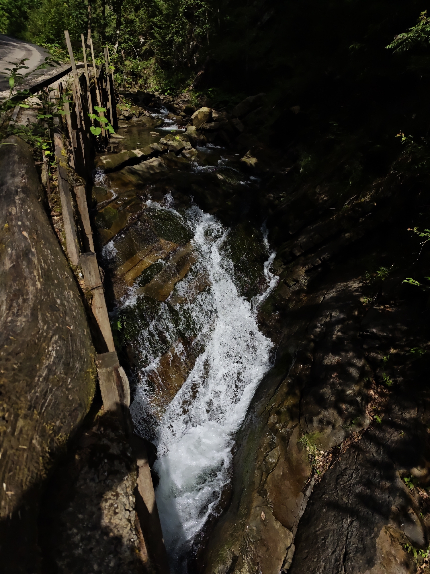 This is a vertical, high-angle shot of a stream flowing through a forested ravine near the city of Tatariv, Ukraine. The stream cascades over rocks and is partially bordered on the left by a wooden retaining wall and a large, rough-hewn log. The water is white and foamy in the foreground where it rushes downwards. The surrounding environment is dense with trees, creating a shadowed, natural scene. A section of a paved road is visible in the upper left corner of the image, adjacent to the wooden structure.