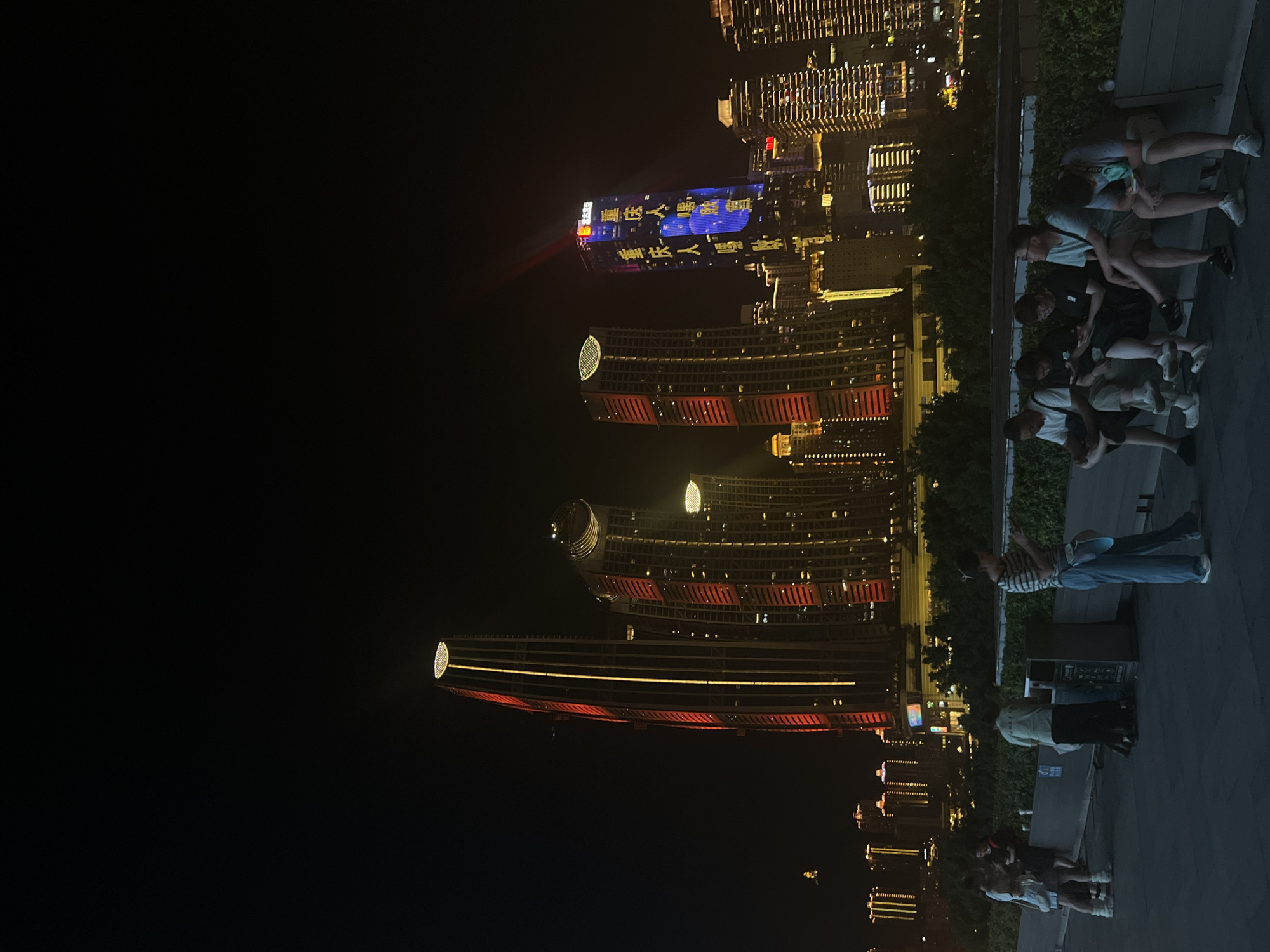 A group of people are gathered on a rooftop at night, overlooking a cityscape. The subjects are a mix of young adults, some sitting and others standing. They appear to be casually dressed, with a few individuals wearing t-shirts and jeans, while others are in shorts. The scene is illuminated by the vibrant lights of the city below and the artificial lights from the buildings.
The backdrop is a sprawling urban landscape, characterized by tall, modern skyscrapers adorned with numerous lights. Some buildings feature distinct architectural elements, such as curved structures and illuminated facades in red and gold. A large digital billboard is visible on one of the buildings, displaying blue text with Chinese characters and a red beam of light emanating from it. The time of day is clearly night, and the weather appears to be clear, allowing for unobstructed views of the city.
The people seem relaxed and engaged in conversation or simply enjoying the view. Some individuals are posing or gesturing, suggesting a casual social gathering. The overall mood is one of enjoyment and appreciation for the urban spectacle. The location is identified as Jiangbeicheng Street in China, which is consistent with the appearance of a modern Chinese cityscape. The presence of streetlights and illuminated buildings confirms the urban setting.