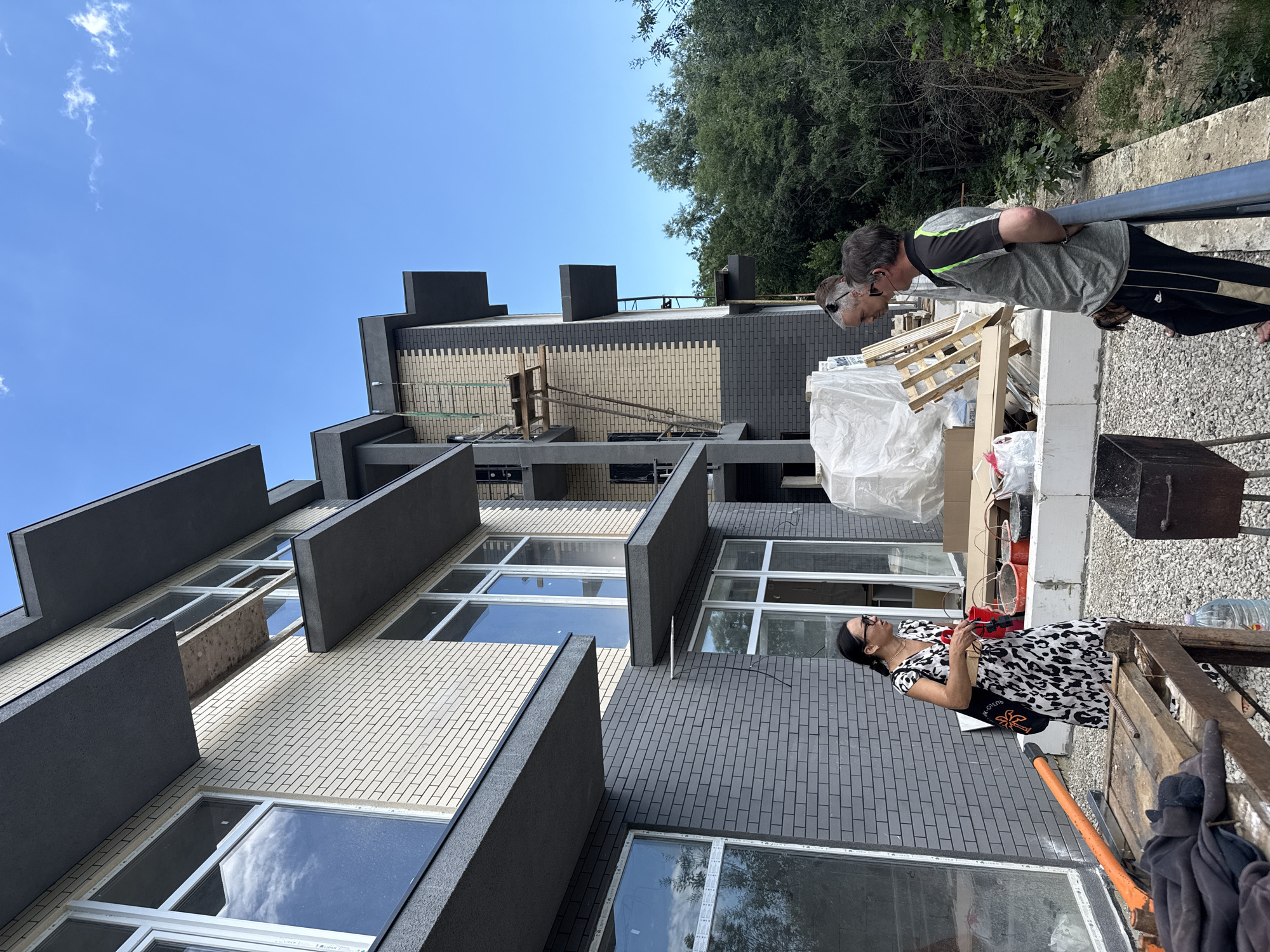 In the city of Yalta, Ukraine, two men and one woman are standing on a balcony of a multi-story building under construction. The building features a light-colored brick facade with dark grey accents and cantilevered balconies. The woman, wearing a black and white patterned dress, stands to the left, facing away from the camera. She is holding a red object and a bag over her shoulder. The two men are positioned to the right, looking towards the building. One man, wearing a grey and green polo shirt and black pants, has his back to the camera. The other man, wearing a light-colored shirt, is partially visible between them. In the foreground, construction materials such as wooden pallets and buckets are visible, along with a portable grill. The background is dominated by lush green trees and a clear blue sky.