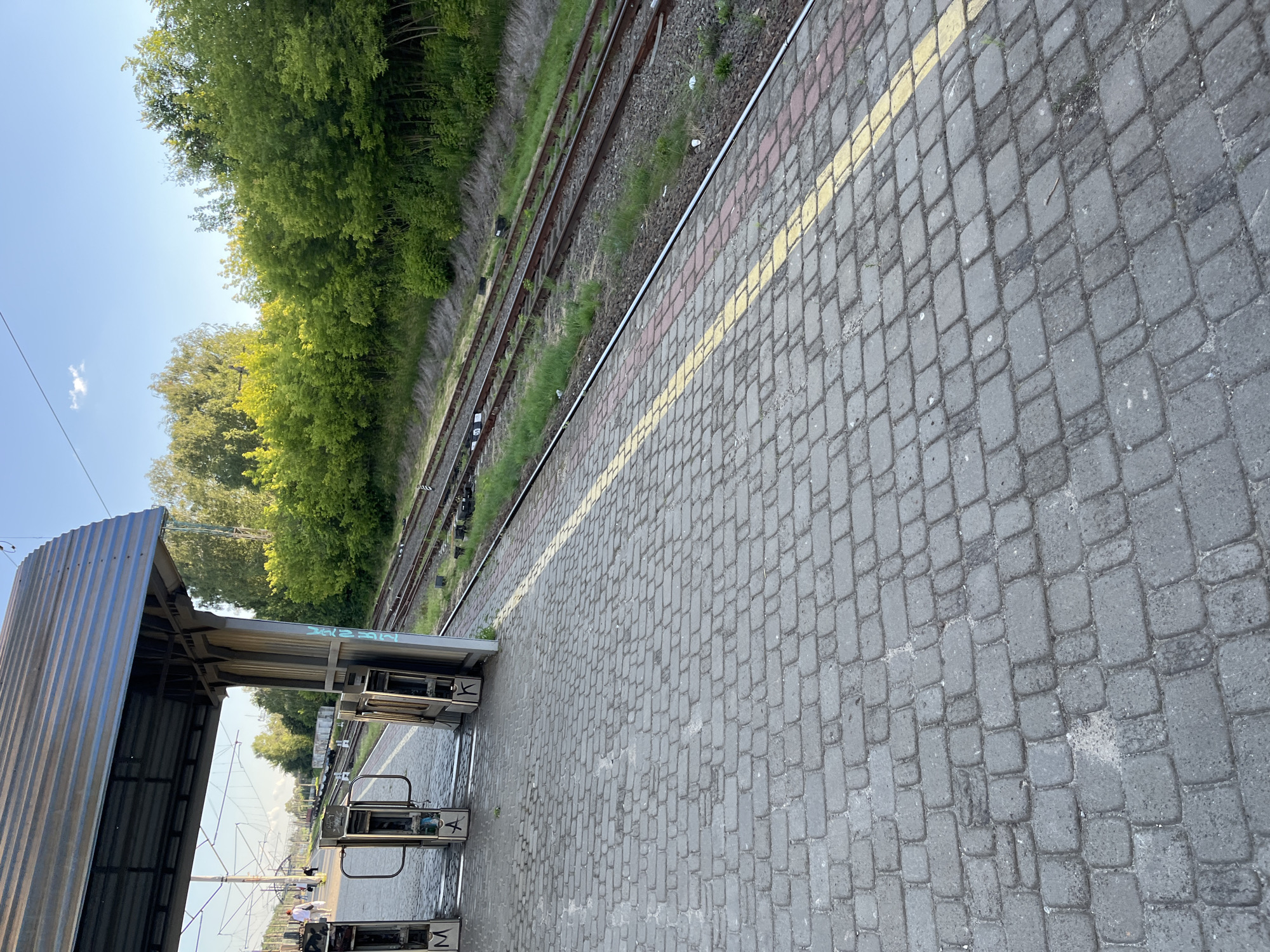 The image depicts a train station platform in Kyiv, Ukraine, on a clear day. The foreground is dominated by a grey, cobblestone platform with a yellow and red striped line running along the edge closest to the train tracks. Several metal turnstiles, some with Cyrillic letters like "M," "A," and "K" spray-painted on them, are visible on the platform. Behind the turnstiles, a covered waiting area with a corrugated metal roof supported by metal pillars can be seen.
In the middle ground, multiple train tracks run parallel to the platform, bordered by a grassy embankment. A line of lush green trees and foliage forms a backdrop along the far side of the tracks, suggesting a natural environment adjacent to the station. The sky above is a clear, pale blue with a single small white cloud. In the distance, a few indistinct figures can be seen walking on the platform, and overhead power lines for the trains are visible. The overall impression is of an active but quiet train station on a pleasant day.