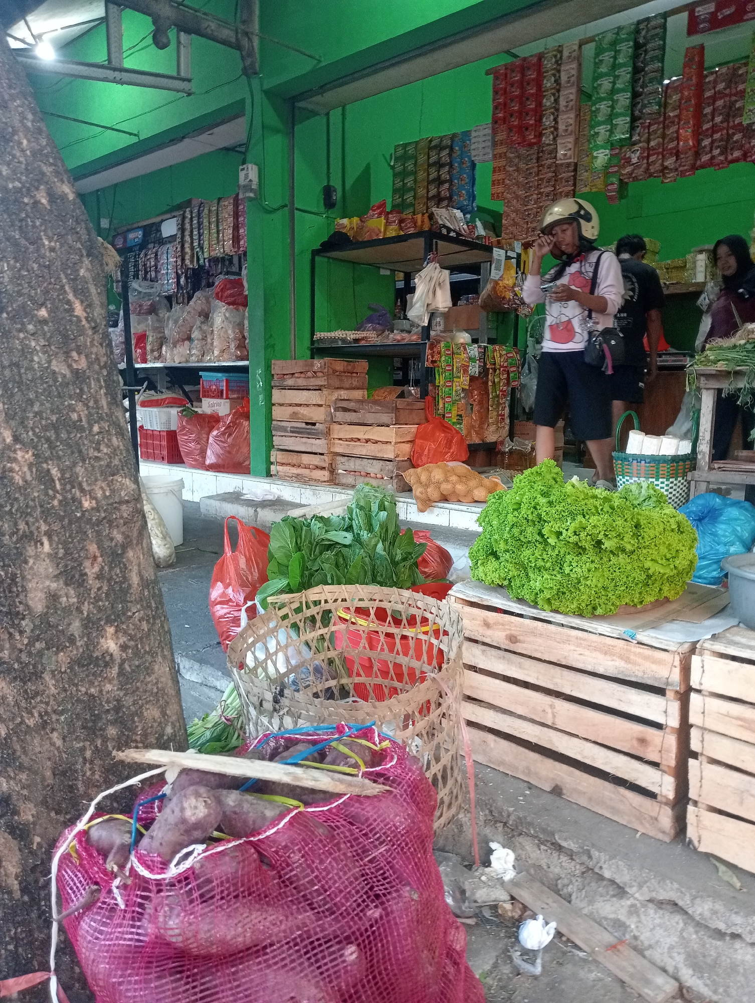 The image depicts an outdoor market scene in Ngawi, Indonesia. In the foreground, a large pile of purple-skinned root vegetables, possibly sweet potatoes, is contained within a magenta mesh bag. To the right, a woven basket sits atop a stack of weathered wooden crates. Adjacent to the basket, a large head of vibrant green lettuce is displayed on a cardboard base, also resting on wooden crates. Behind the lettuce, a blue plastic bag and a portion of a gray plastic tub are visible. A collection of leafy green vegetables, likely bok choy or similar, is arranged in front of a red plastic basket. Further back, several red plastic bags and white plastic crates are stacked. To the left, a substantial tree trunk with rough bark dominates the frame. In the mid-ground, a woman wearing a yellow helmet and a light-colored shirt with a cartoon design is looking at her phone. Another person, partially obscured, stands behind her. The background reveals shelves stocked with packaged goods, including snacks and instant noodle packets, displayed against a bright green wall. The overall scene suggests a bustling local market.