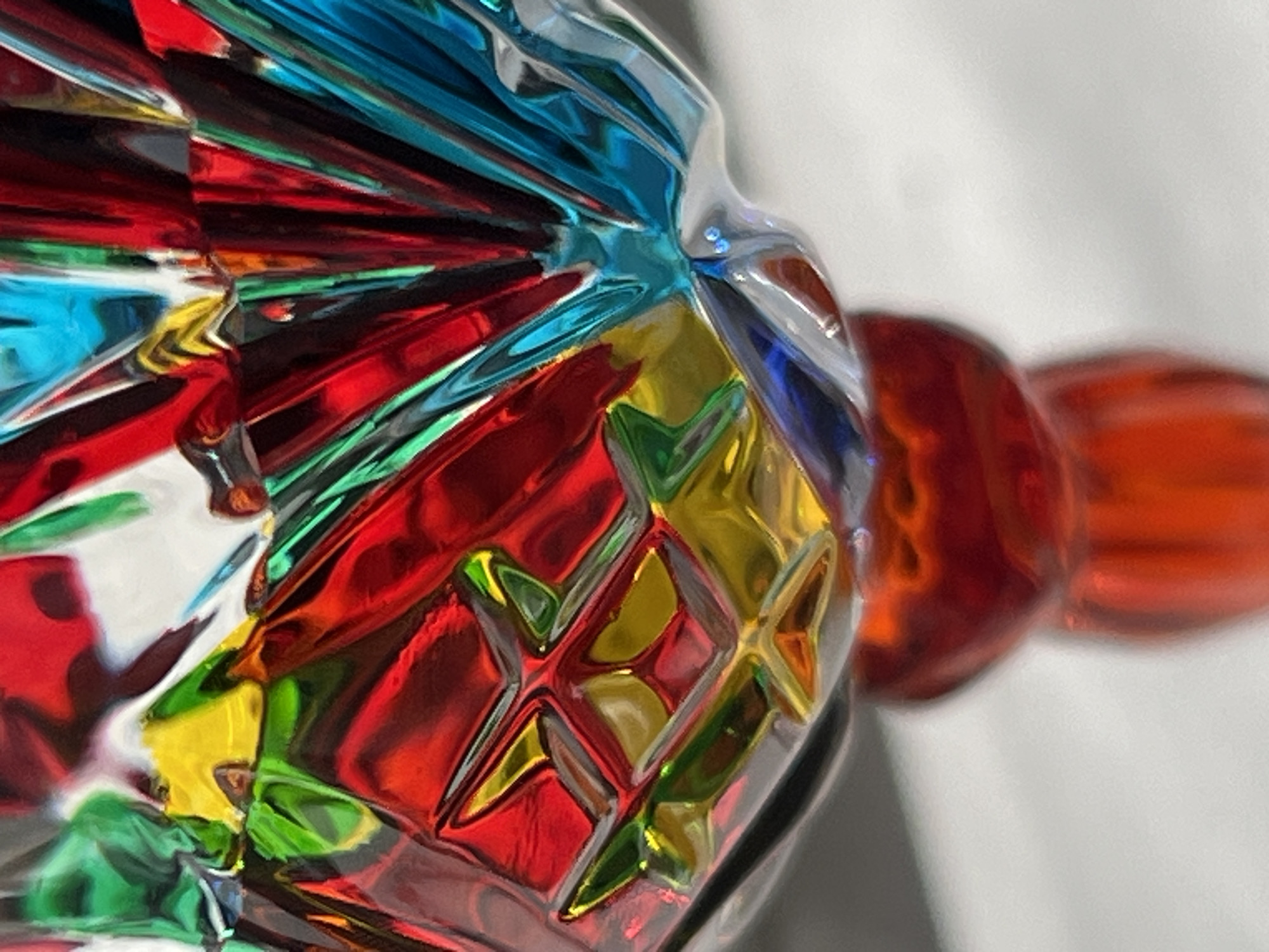 This is a close-up, abstract photograph of a multi-colored glass object. The object appears to be faceted, with many sharp, reflective surfaces. The colors are vibrant and include shades of red, blue, yellow, and green, with some silver-toned areas as well. The light catches the facets, creating bright highlights and deep shadows, giving the object a dynamic and almost crystalline appearance. Towards the right side of the image, there are smooth, rounded forms of a deep orange or red color, suggesting they are either part of the same object or a separate element positioned behind it. The background is out of focus and appears to be a plain, light gray surface, suggesting a studio setting or a neutral environment. There is no discernible activity or event taking place; the focus is purely on the texture, color, and reflective properties of the glass. No people, text, or specific location cues are visible. The overall impression is one of artistic abstraction, highlighting the beauty of light interacting with a complex glass form.