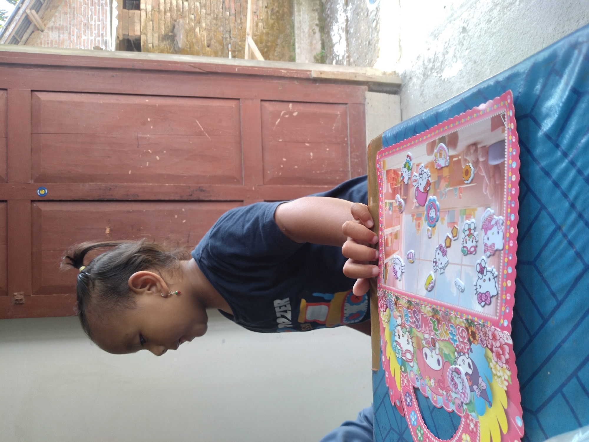 A young Indonesian girl, with her hair pulled into a ponytail and wearing an earring, is shown from the side. She is wearing a dark blue t-shirt with a graphic print that includes the word "DOZER". Her right hand is resting on a sticker book on a blue table. The sticker book is pink and features various cartoon characters, including Hello Kitty. The background shows a wooden door and a brick wall, suggesting an outdoor or semi-outdoor setting. The image is likely taken in the city of Pandak, Indonesia, given the context provided.