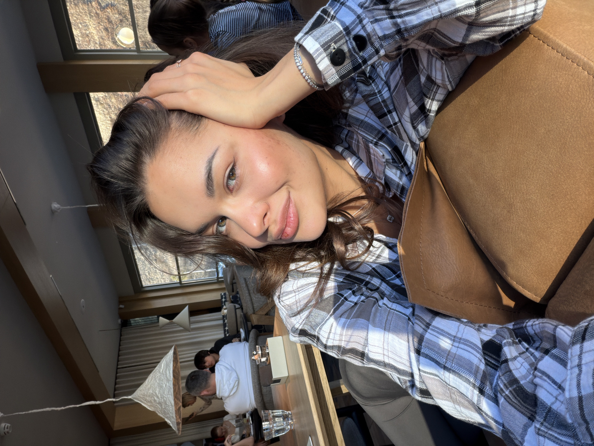 The photograph shows a young woman with brown hair and green eyes indoors. She is wearing a plaid shirt and a bracelet. Her hand is resting on her head. In the background, there are other people at tables in what appears to be a restaurant. A geometric lampshade hangs from the ceiling. The location could be inferred to be in Krasnoyarsk, Russia based on prompt context.