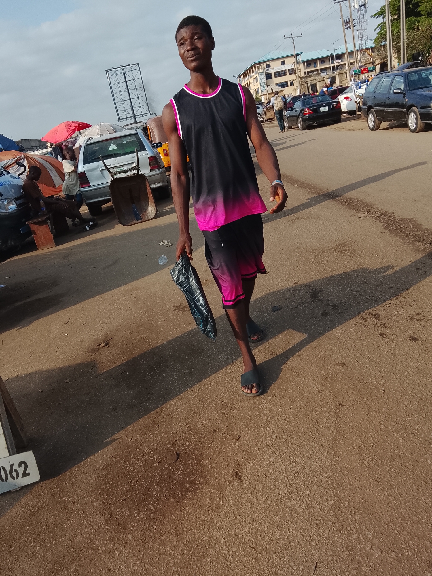 A young man walks towards the camera on a paved street. He is wearing a black tank top with pink trim and black-to-pink gradient shorts, and blue sandals. He carries a dark, bundled object in his left hand. His shadow stretches behind him on the road. Behind him, there is a bustling street scene including cars, pedestrians, small shops, and a multi-story building in the distance. The scene appears to be in a market area in Abuja, Nigeria. There are several parked cars and people walking about. A billboard frame stands near a white car with a wheelbarrow nearby.