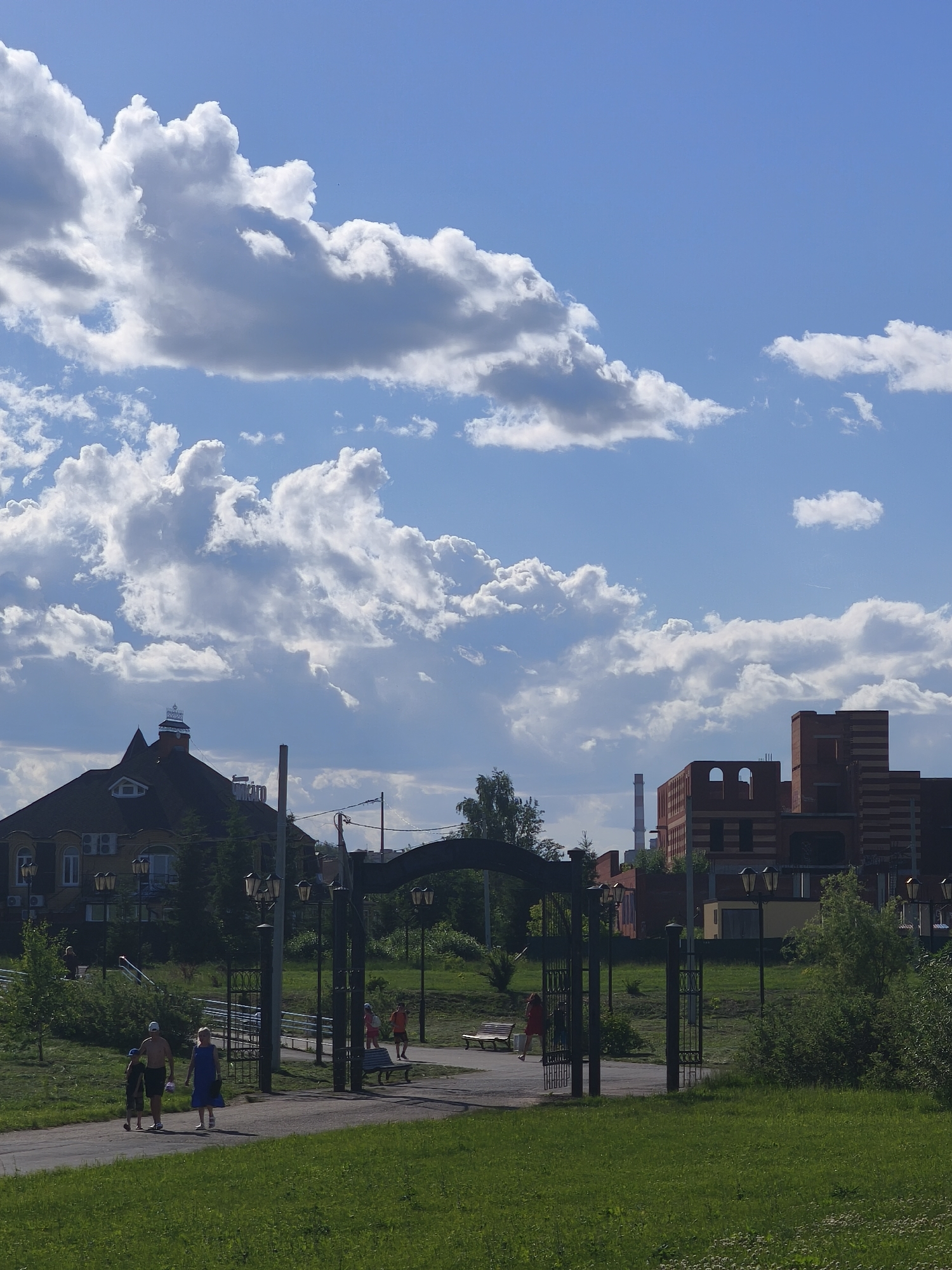The image depicts a daytime scene in a park-like setting, likely in Yoshkar-Ola, Russia. A paved path winds through a grassy area, bordered by trees and bushes. Several people are present, including a family walking together on the path, and other individuals scattered in the background.
In the foreground, a family consisting of two adults and two children are walking away from the camera on the path. The adult male is wearing shorts and is shirtless, while the adult female is wearing a blue dress. The two children are also wearing shorts.
Further along the path, several other individuals are visible. One person in a red top and dark shorts is walking away from the camera. Another person in an orange top and dark shorts is also walking away from the camera. Two benches are situated along the path.
An ornate metal archway with decorative gates is a prominent feature in the mid-ground, framing the path. Several lampposts with multiple lights are positioned along the path and near the archway.
In the background, a large, multi-story building with a dark roof and dormer windows is visible on the left. To the right, a modern, multi-story brick building with arched windows is present. A tall, slender industrial chimney or tower is also visible in the distance. The sky above is a vibrant blue with large, fluffy white clouds. The overall impression is a pleasant, sunny day in a public outdoor space.