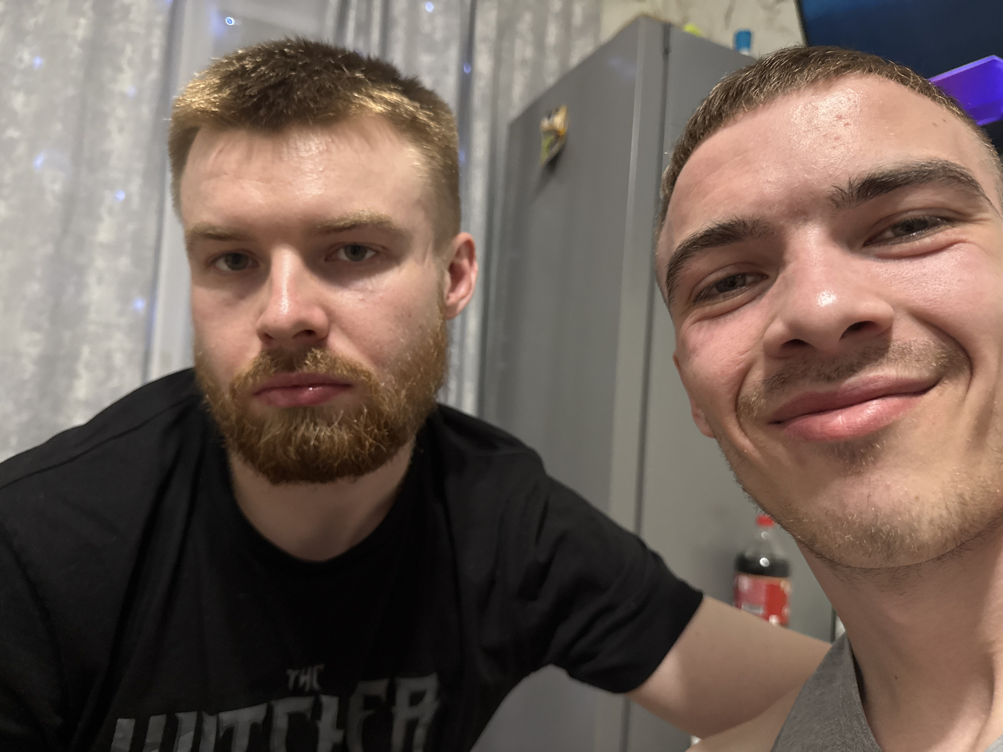 Two men are indoors taking a selfie. The man on the left has short, light brown hair and a full, ginger-brown beard. He is wearing a black t-shirt. The man on the right has short, dark brown hair and a light stubble beard. He is wearing a gray tank top. Behind them is a gray refrigerator, a bottle of Coca-Cola, and white curtains with small lights. The location is in a home.