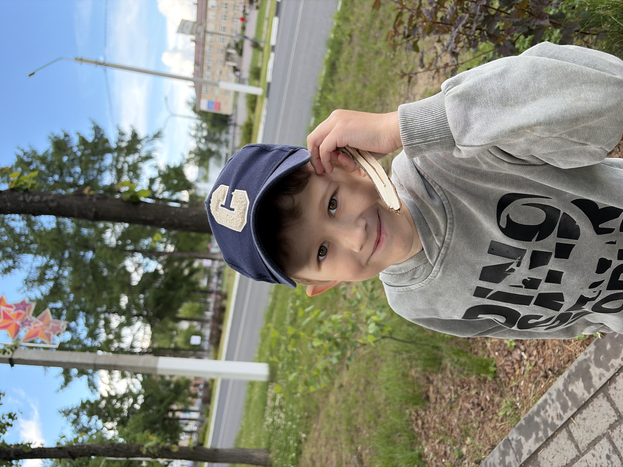 The image, correctly oriented, features a young boy standing outdoors in what appears to be Chaikovsky, Russia, on a bright, sunny day. The boy is positioned slightly to the right of the frame, looking directly at the camera with a soft smile. He has light skin, dark hair, and light-colored eyes. He wears a navy blue baseball cap adorned with a large, beige letter "G" on the front, and a light grey long-sleeved sweatshirt with stylized black text or a design visible across the chest. His right hand is raised, holding a light-colored piece of wood or bark near his face.
The background shows an urban or suburban environment. To the boy's left, a row of tall trees with green foliage lines a paved road. A clear blue sky with sparse white clouds dominates the upper portion of the scene. In the distance, a multi-story, light-colored building can be seen, along with several streetlights or utility poles. On the far left, a decorative element composed of orange and red stars is attached to a pole. The foreground consists of a grassy area and a paved sidewalk or path, which has visible wet patches, suggesting recent rain. The overall scene is a casual outdoor portrait.